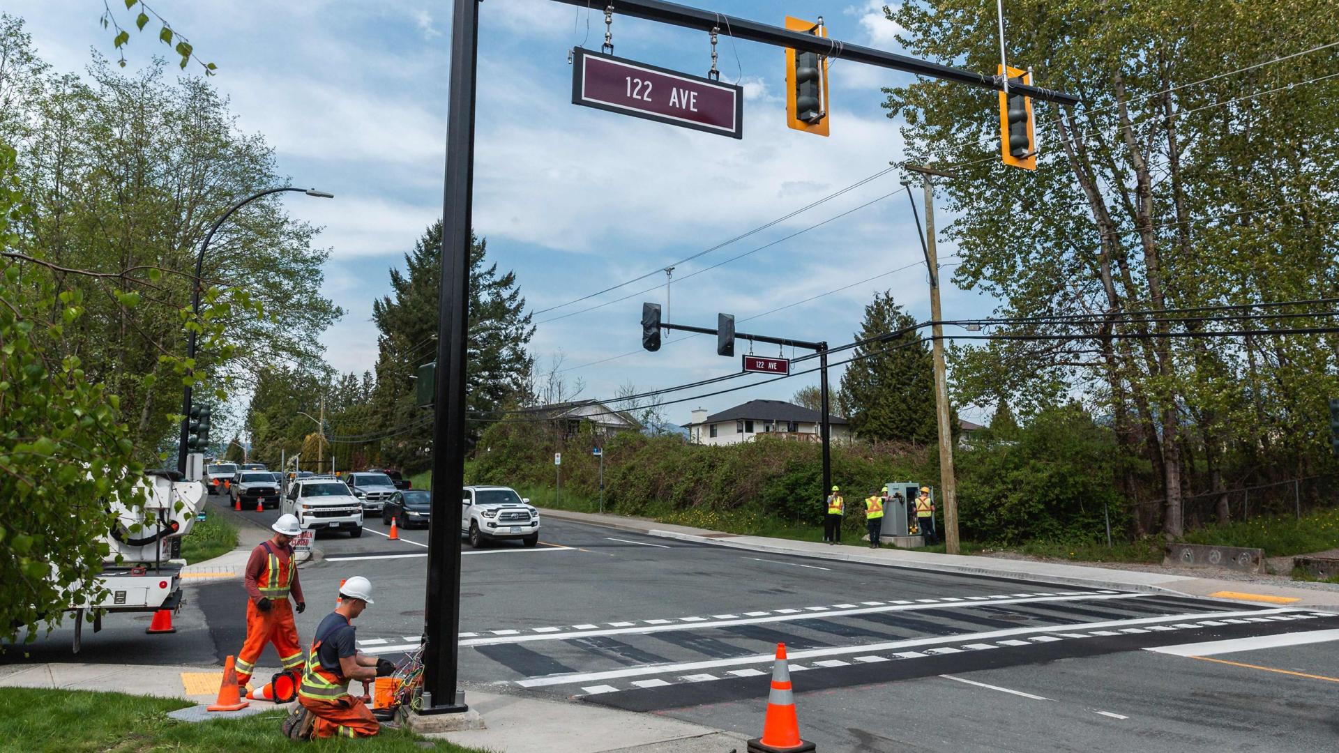 Two workers work on the post suspending a traffic light over a three-way intersection. A freshly painted crosswalk runs under the traffic lights. 