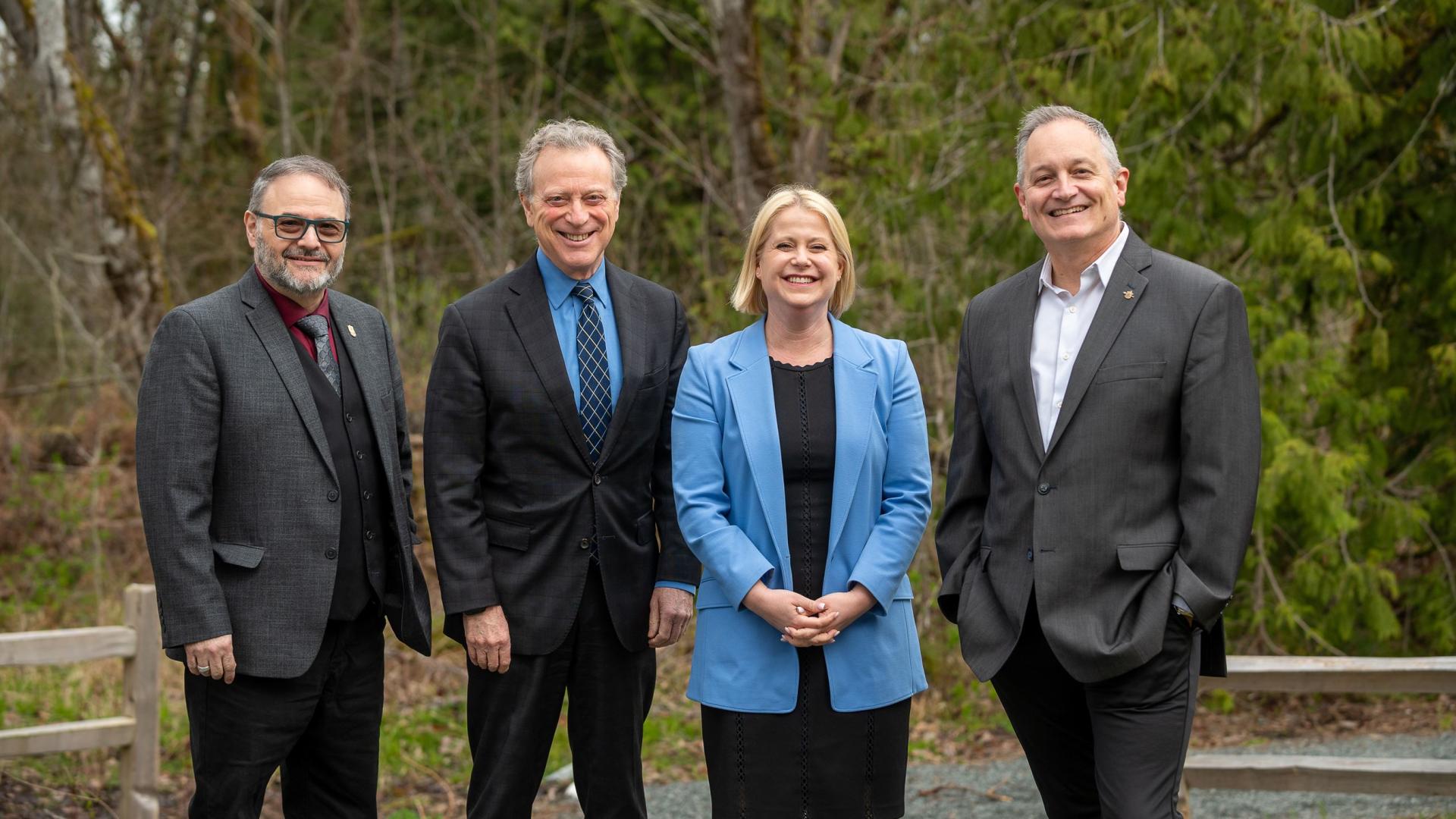 3 men and a women stand in a line, smiling for a photo outdoors.
