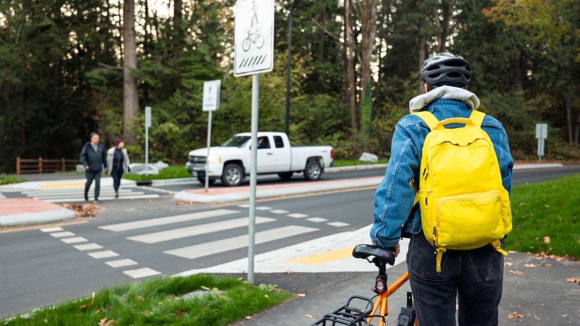 A person walking with a bike approaches the crosswalk at a round about. Across the road, a couple crosses as a truck waits for them.