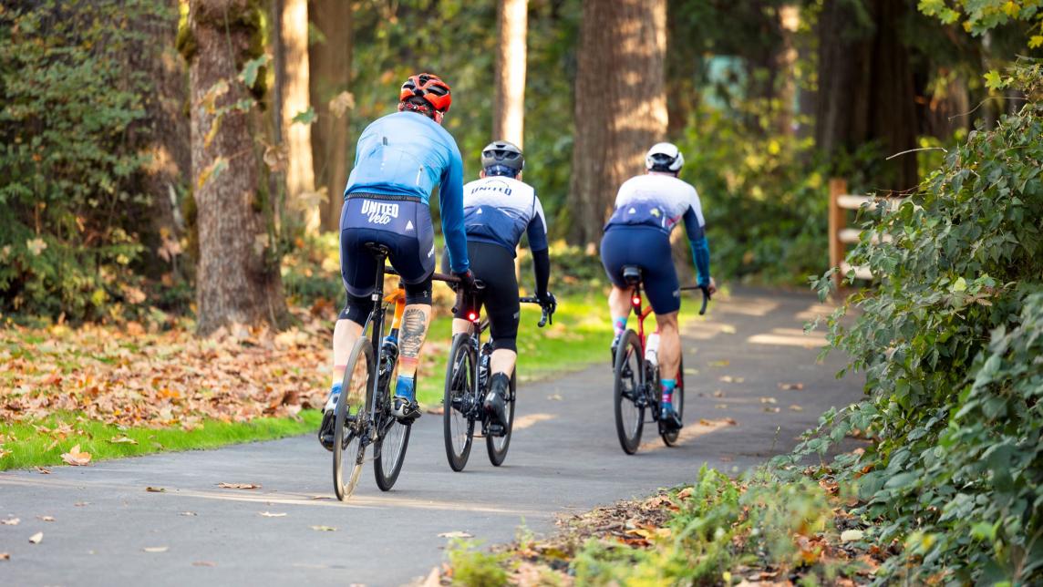Three bikers in full bike gear bike in a line on a paved path.