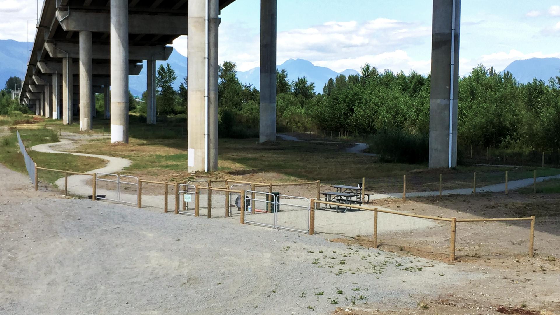 Underneath an overpass on the other side of a gravel parking lot is a fenced-in grassy area with a winding paved path.