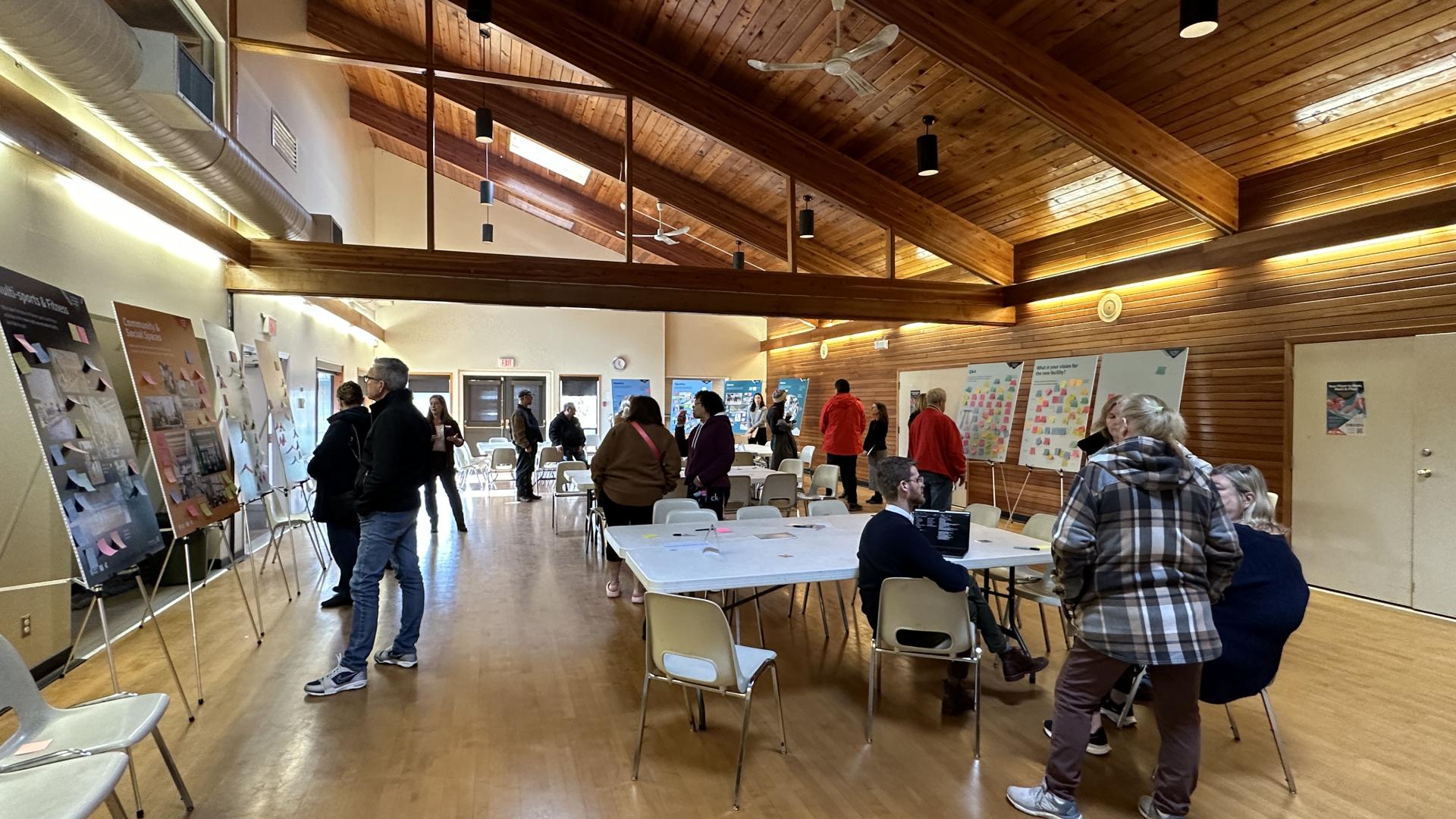 A crowd sits at white plastic tables and surveys posterboards within a gym-like room. The ceiling is slanted and forms a peak against on wall and the floors are a polished wood. 