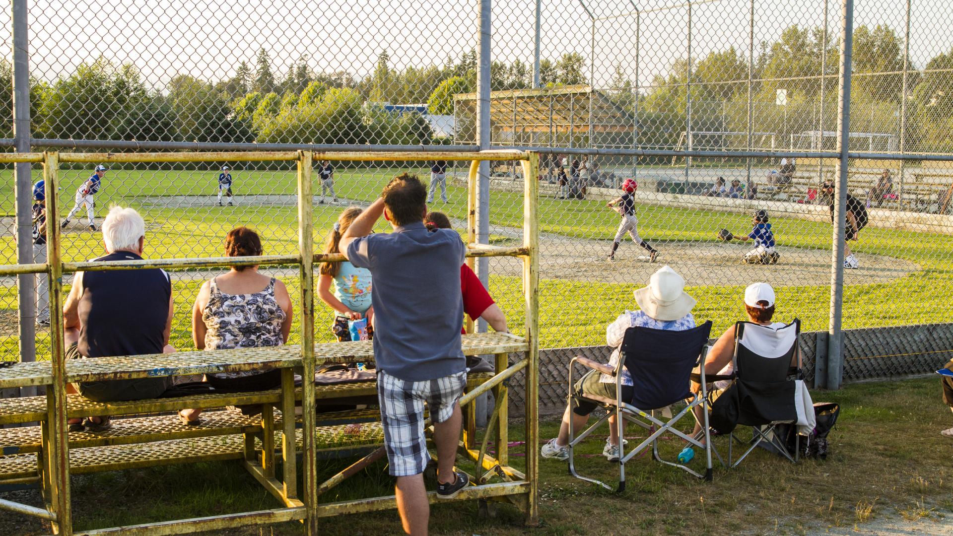 A crowd of people watch a baseball game from the other side of the fence on bleachers and lawn chairs at the Albion Sports Complex.