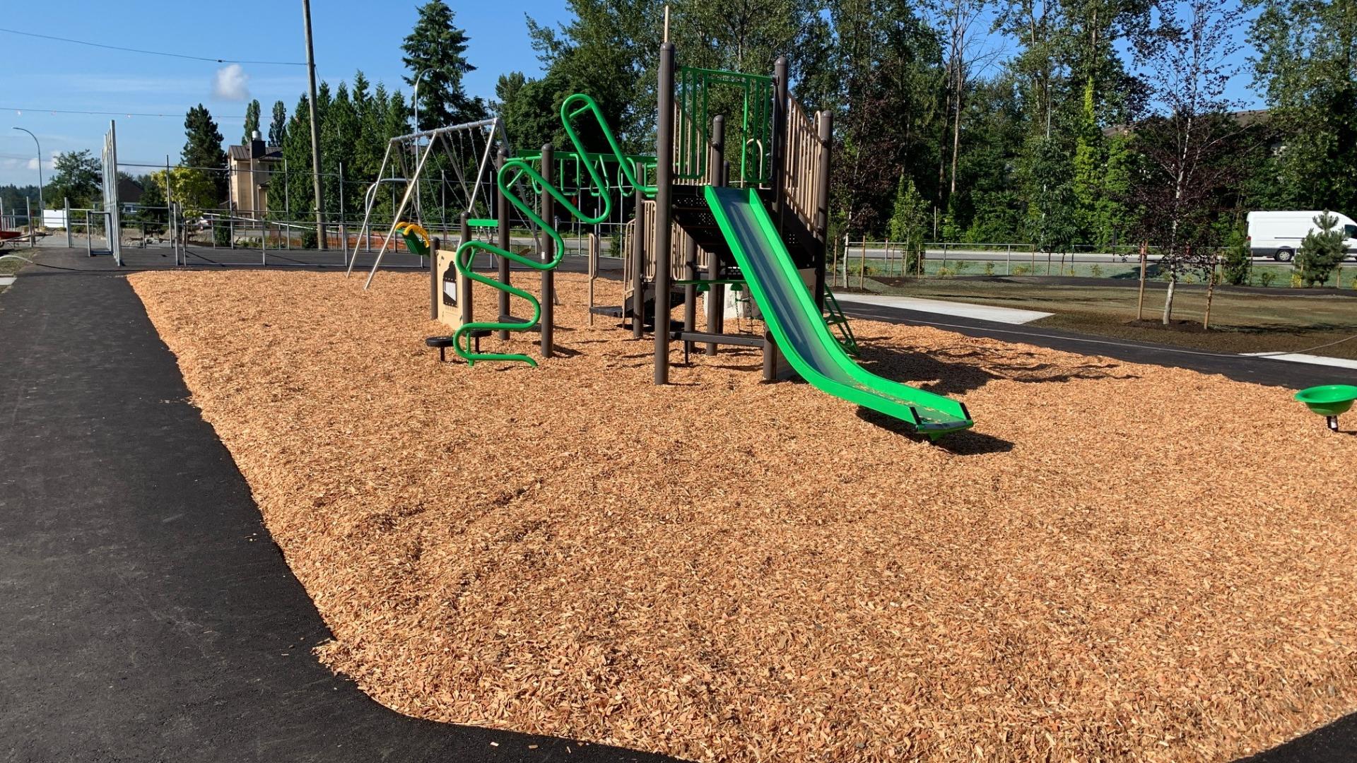 A black and green playground on mulch surrounded by a wide, paved path. On the far side there are benches.
