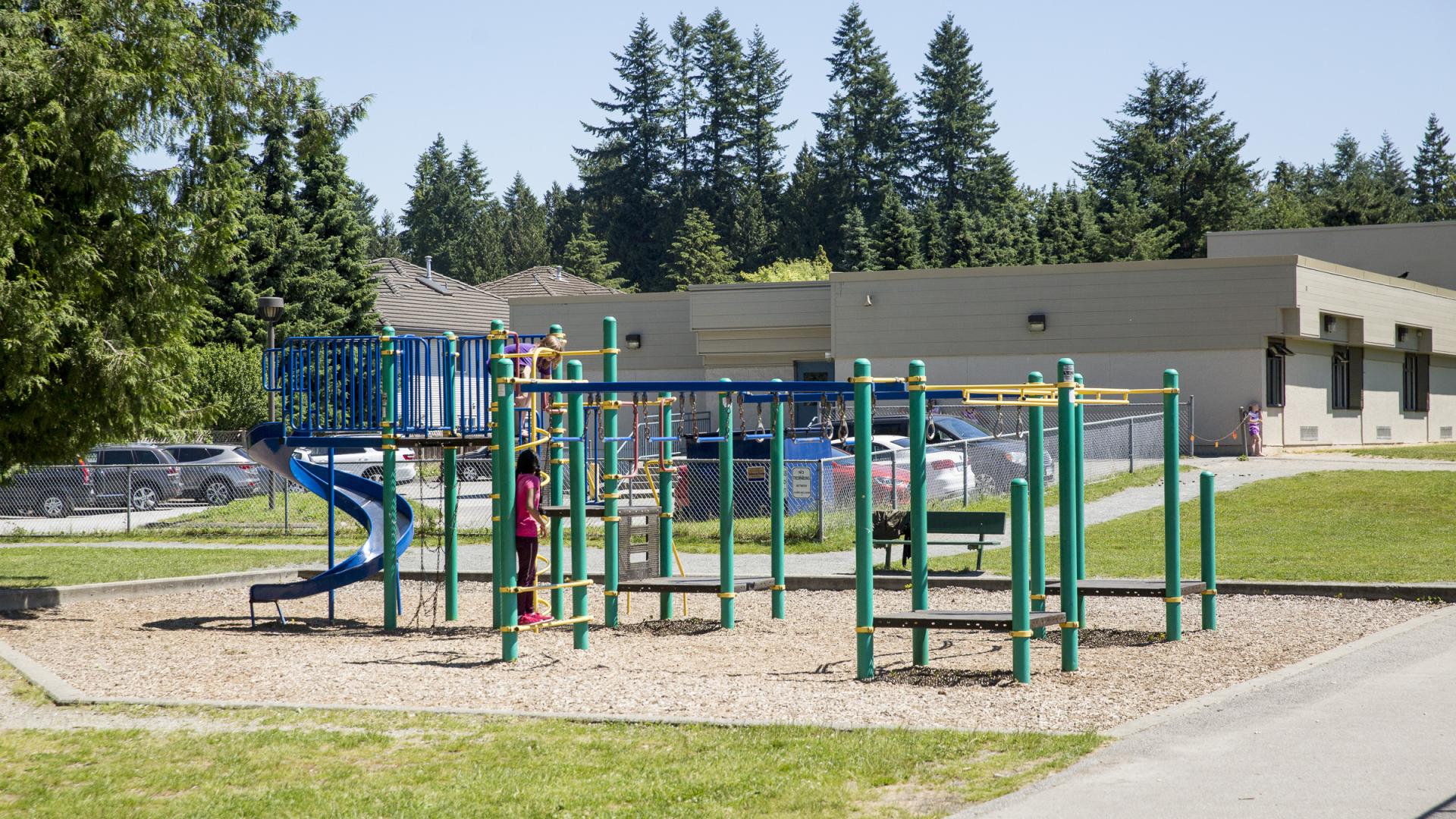 A green playground with a large twisty slide sits alongside a paved pathway in a grassy field with a barch mulch play area.
