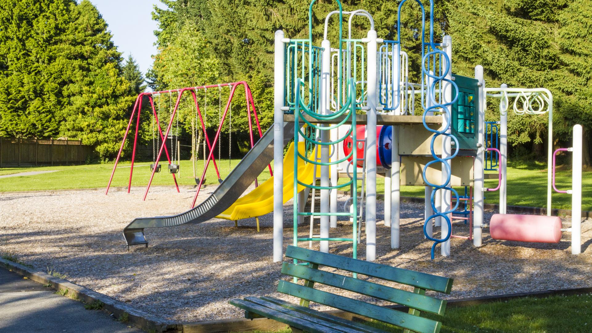A brightly coloured playground behind a small bench and in front of a swing set.