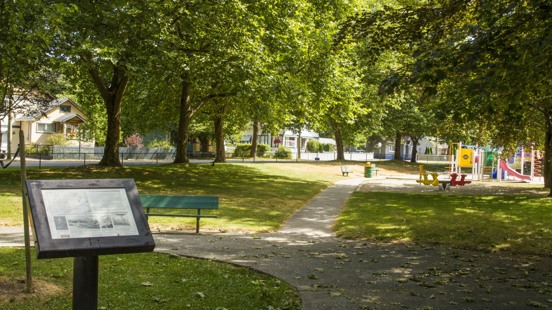 A well shaded path runs past a small information sign and toward a colourful playground..