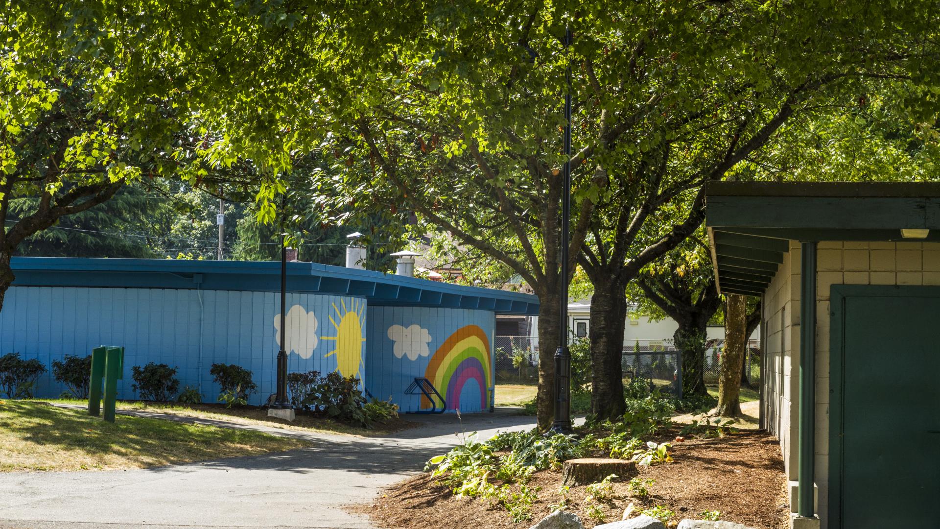 A bright blue building with a sun, clouds and a rainbow painted on it sits next to a tree lined paved walkway.