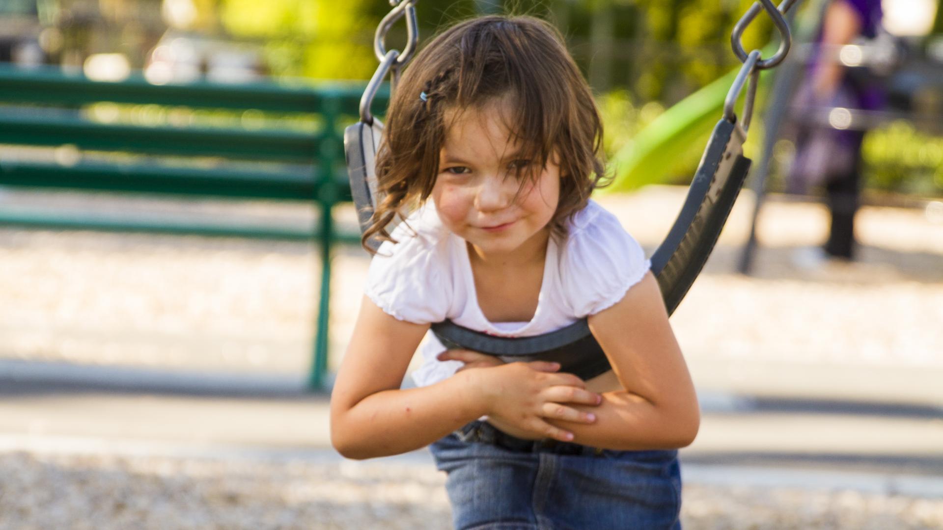 A young girl swings stomach down on a swing. The mulch under the swing has been brushed aside exposing the vinyl below. 