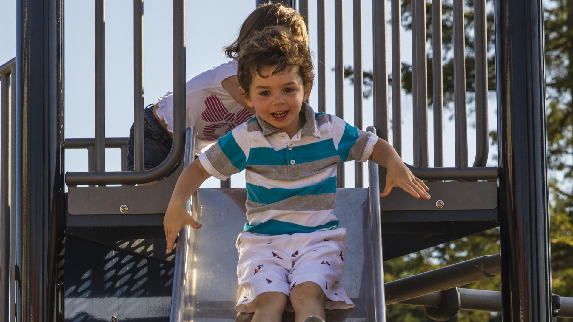A toddler age boy slides down a metallic slide.