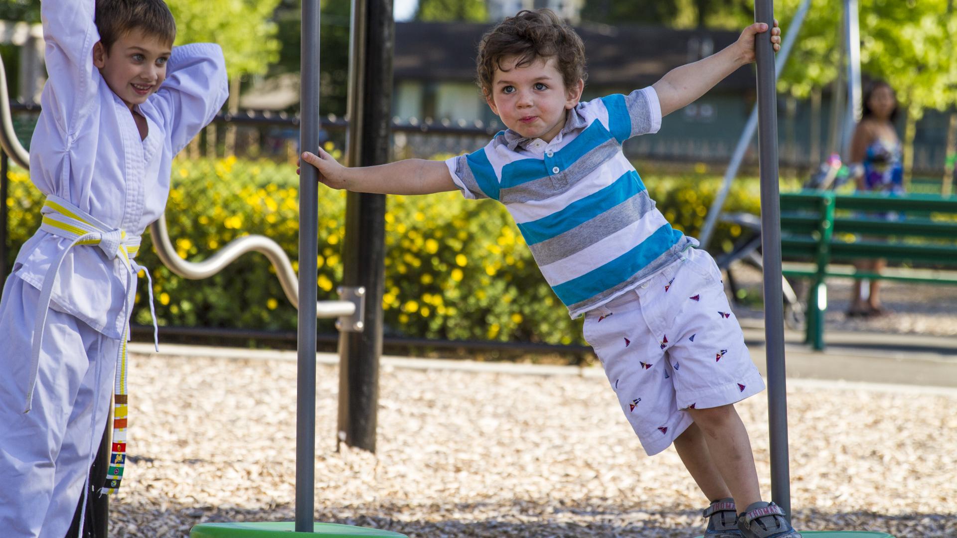 A toddle age boy and a young boy in a Gi hang off of floating stone style playground equipment. 
