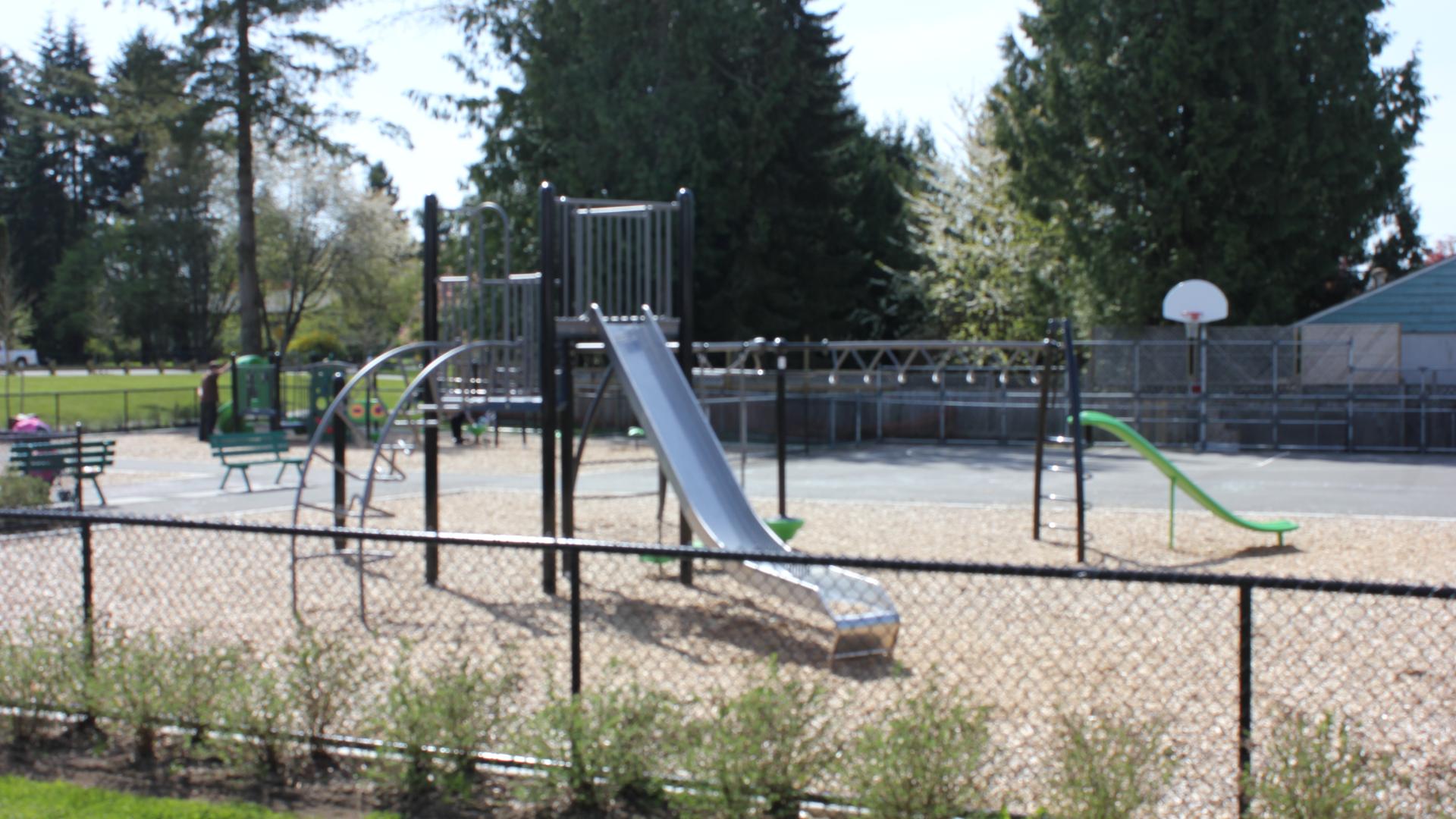 A fenced in playground with two free standing metallic slides. 
