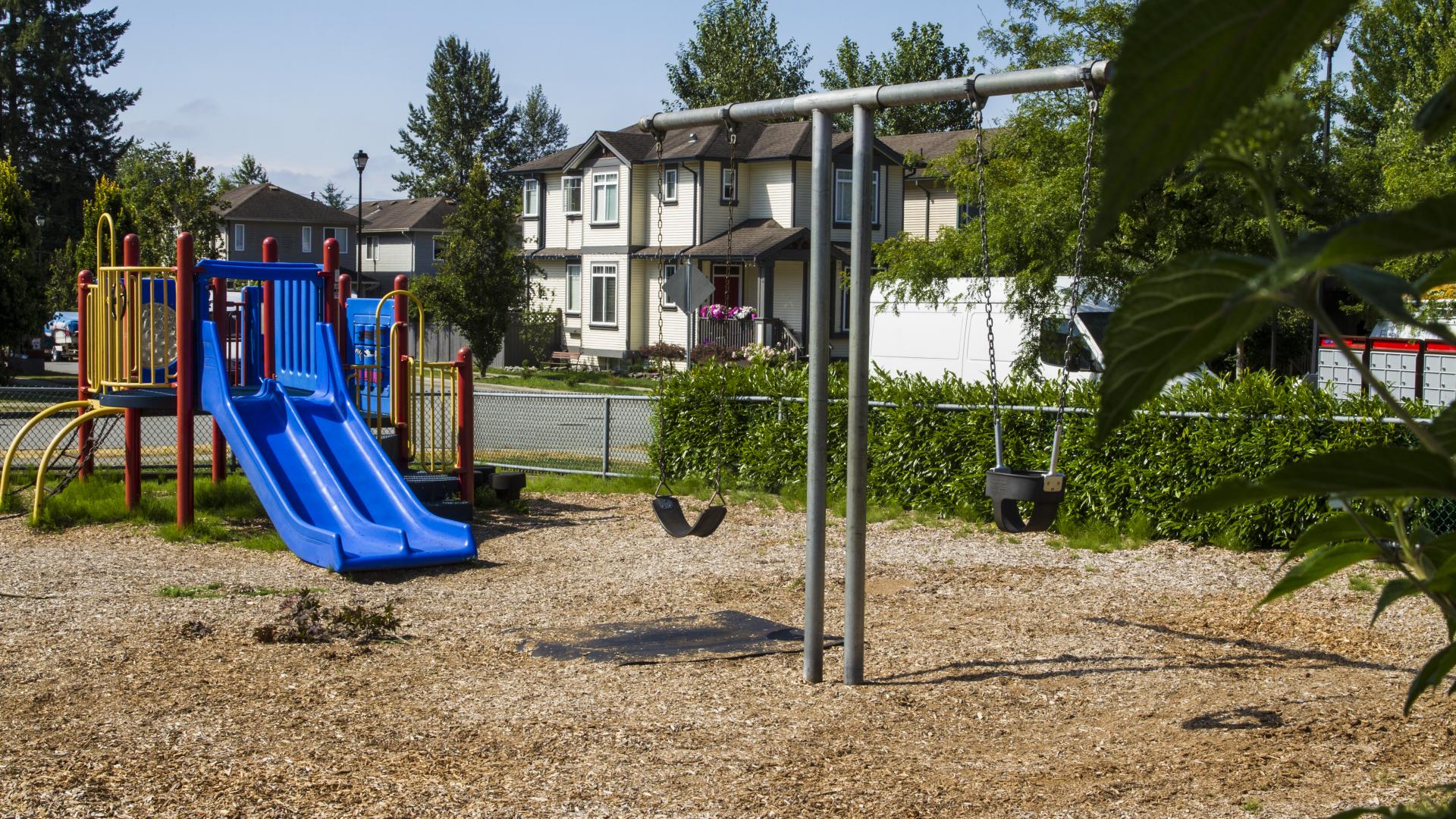 A playground on a mulch bed behind a T-shaped swing set. There are residential houses nearby, backing into the park.