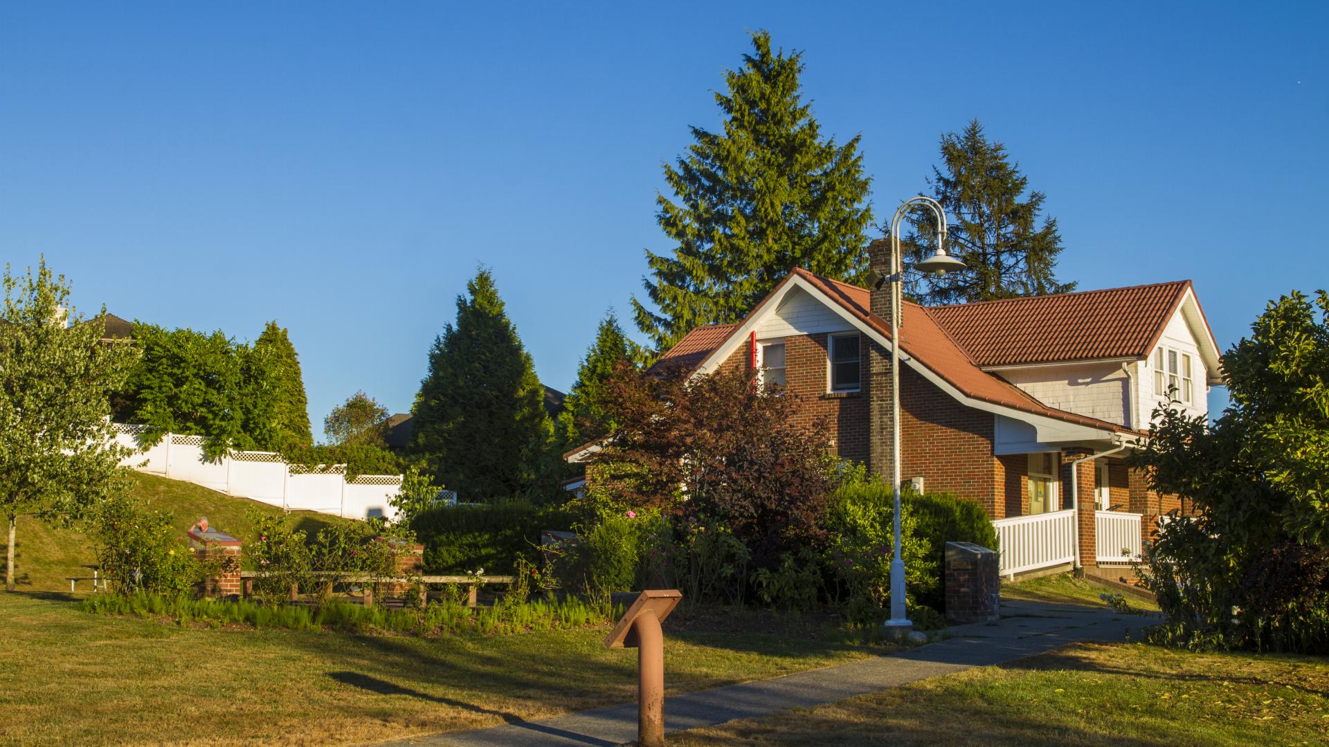 A paved path with a small sign next to it leads to a medium sized hours surround by many trees.