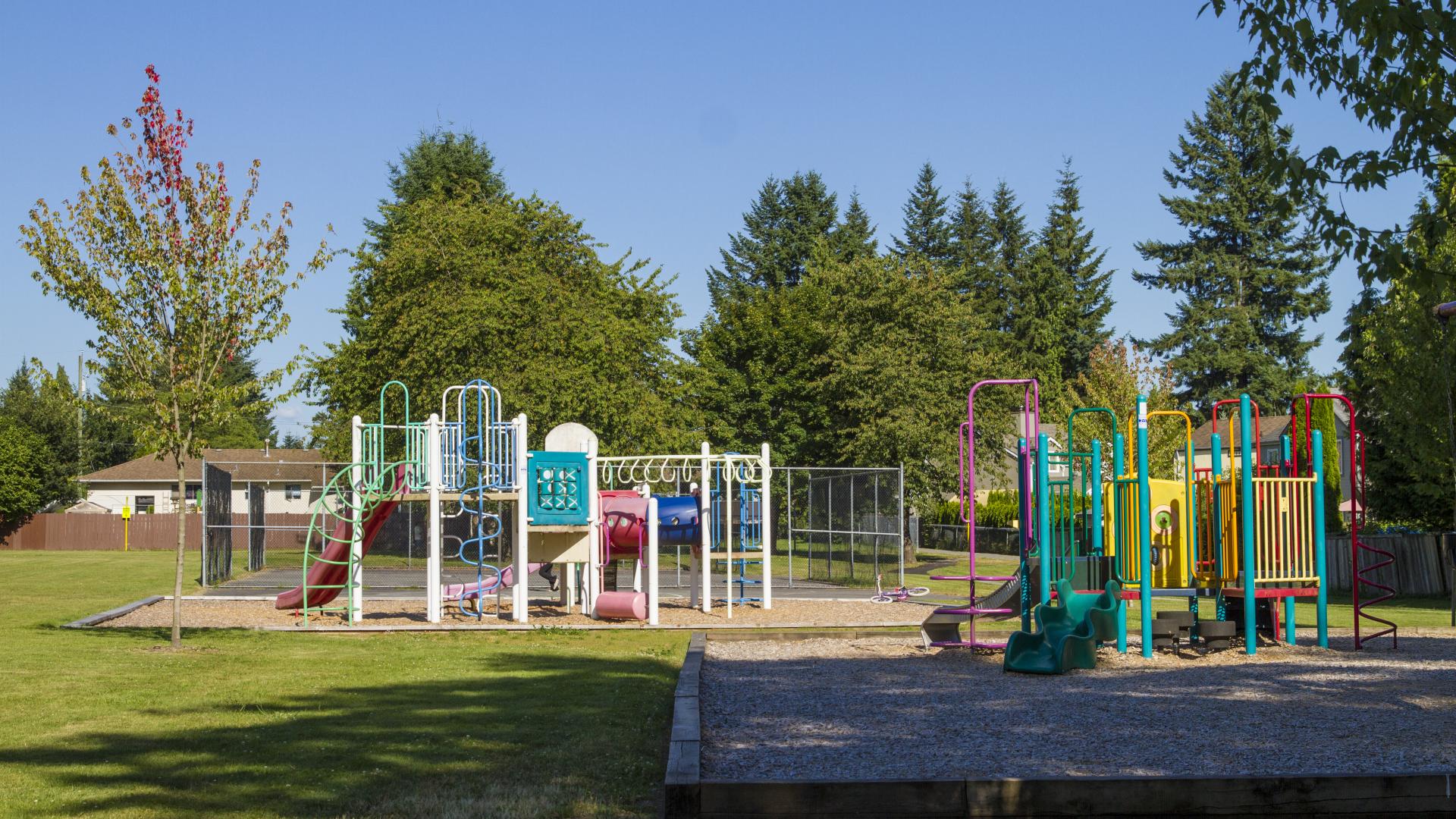 Two bright coloured playgrounds. The shade from near by trees in creeping up on one, while the other is in full sun.