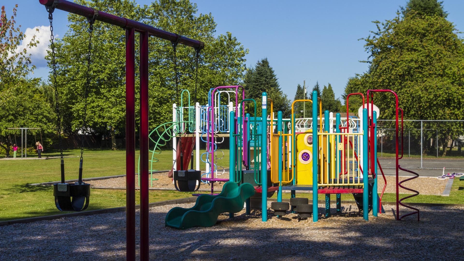 A t-bar shaped swing set and two bright playgrounds in a large, sunny grassy field.