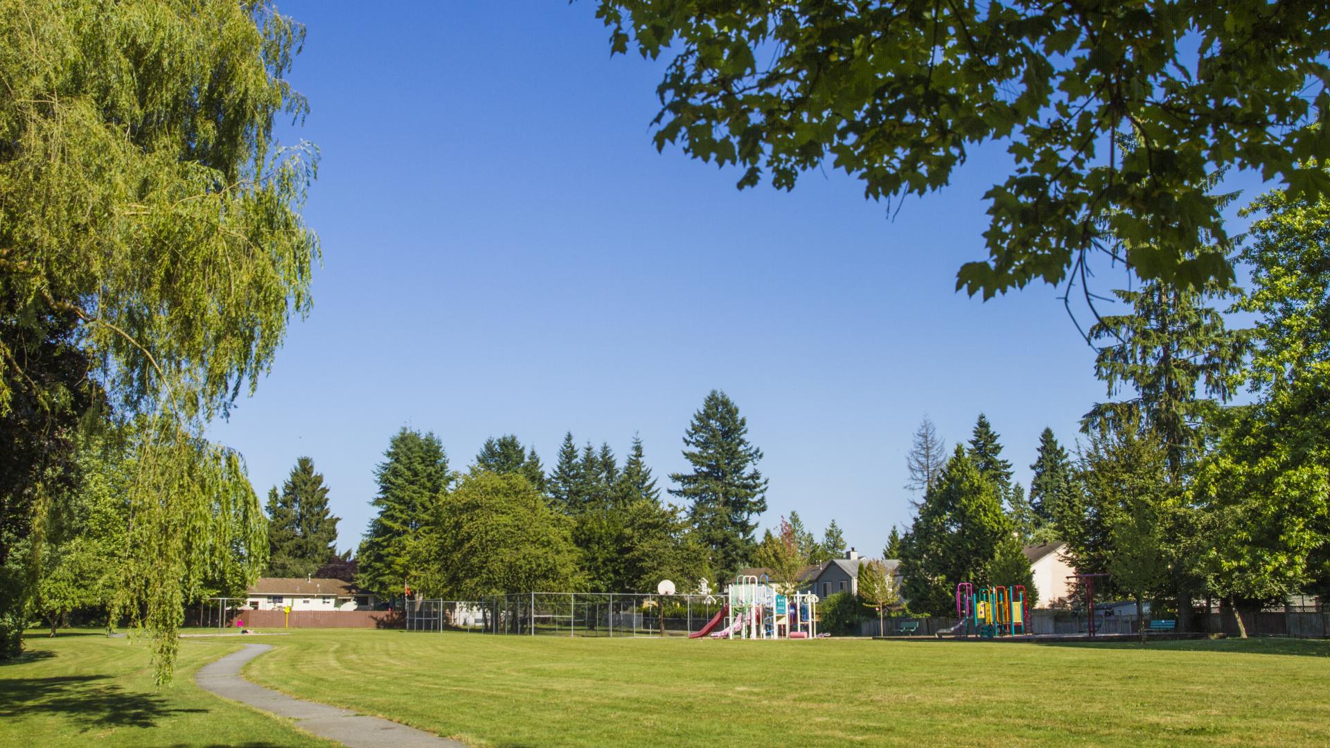 A wide open grass field. A paved path arcs through the centre of it and tree's surround the perimeter. 