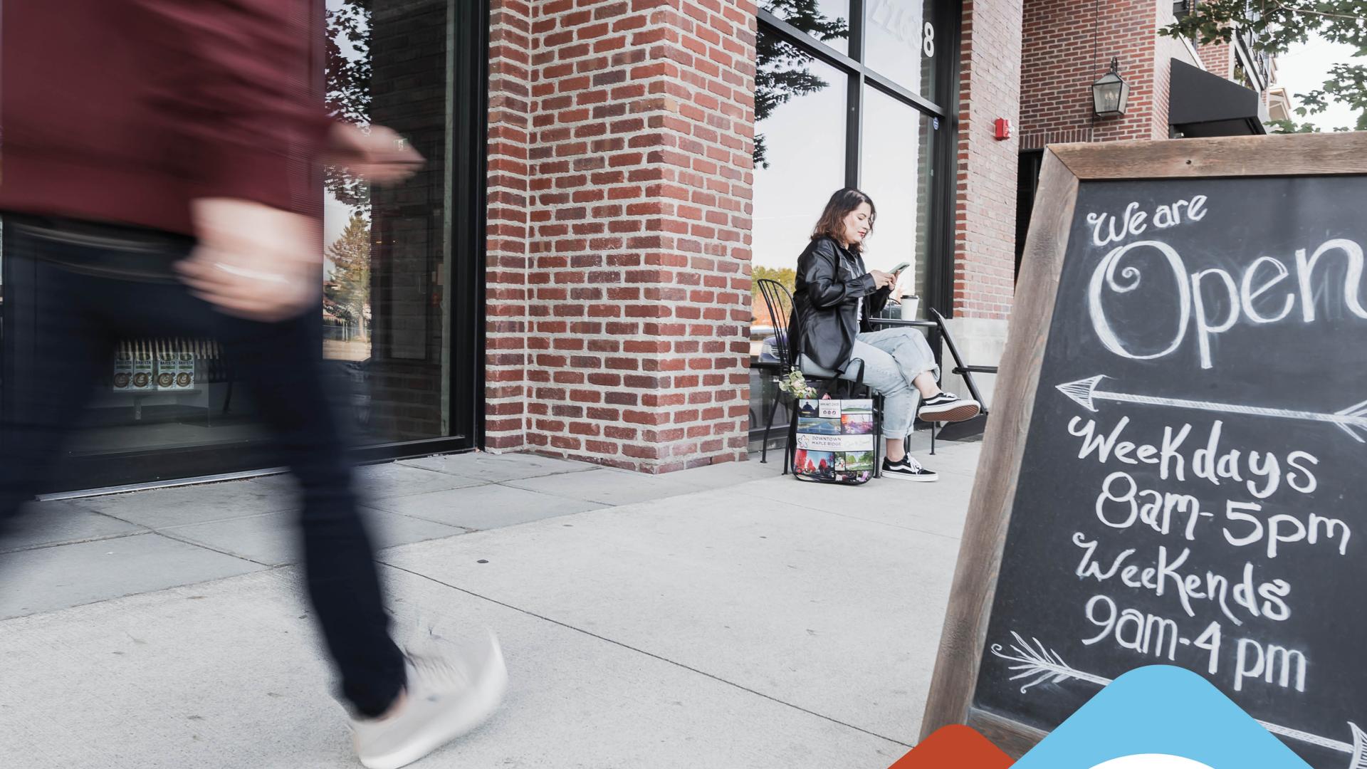 A person walks by a chalkboard "Open" sign for a store, while another person rests on a ledge checking their phone above a blue portion with text that reads "News Release Free Buy Local Workshop for Maple Ridge Businesses on April 8".