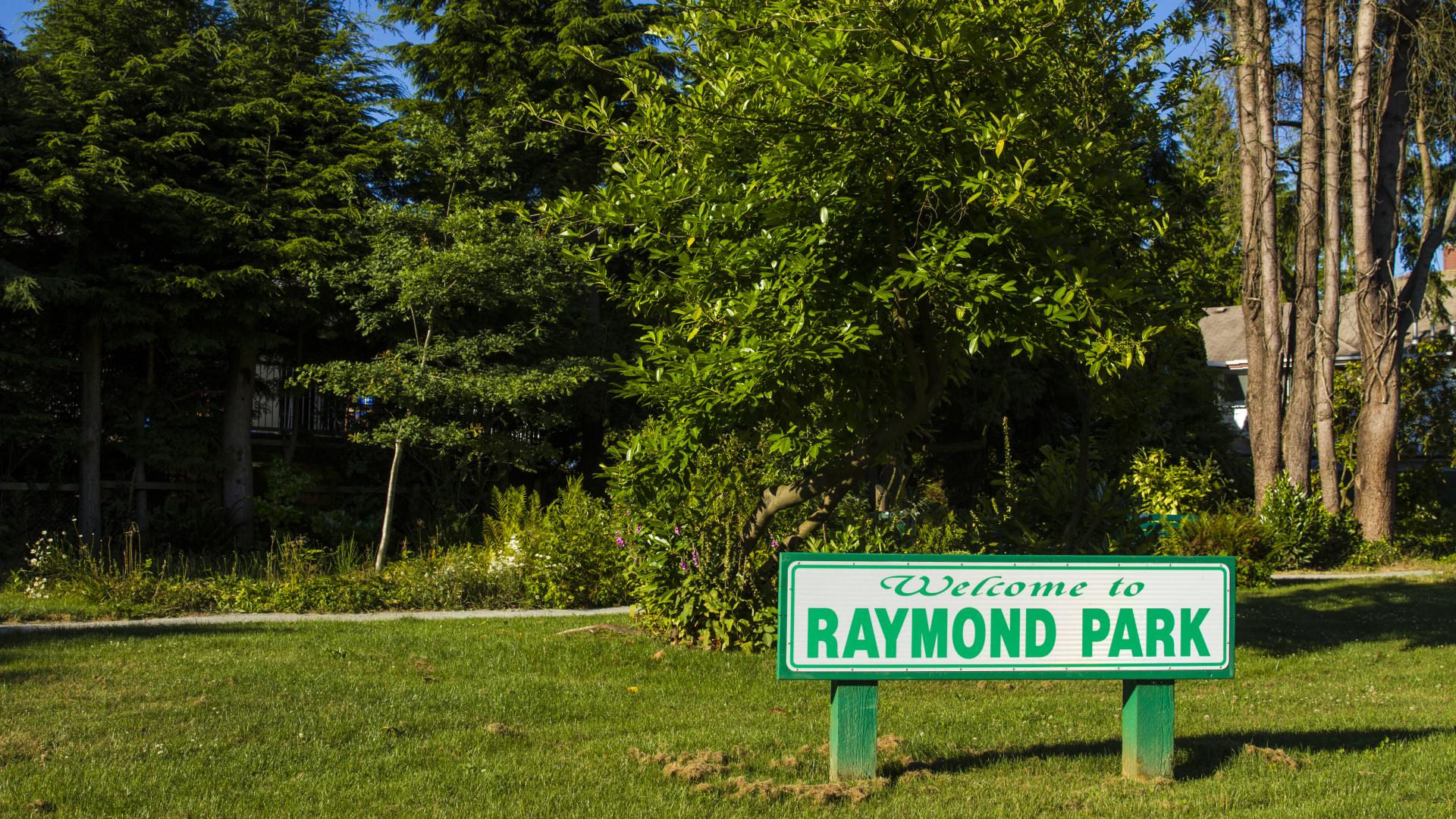 The Raymond Park sign on a patch of grass in front of a trail.