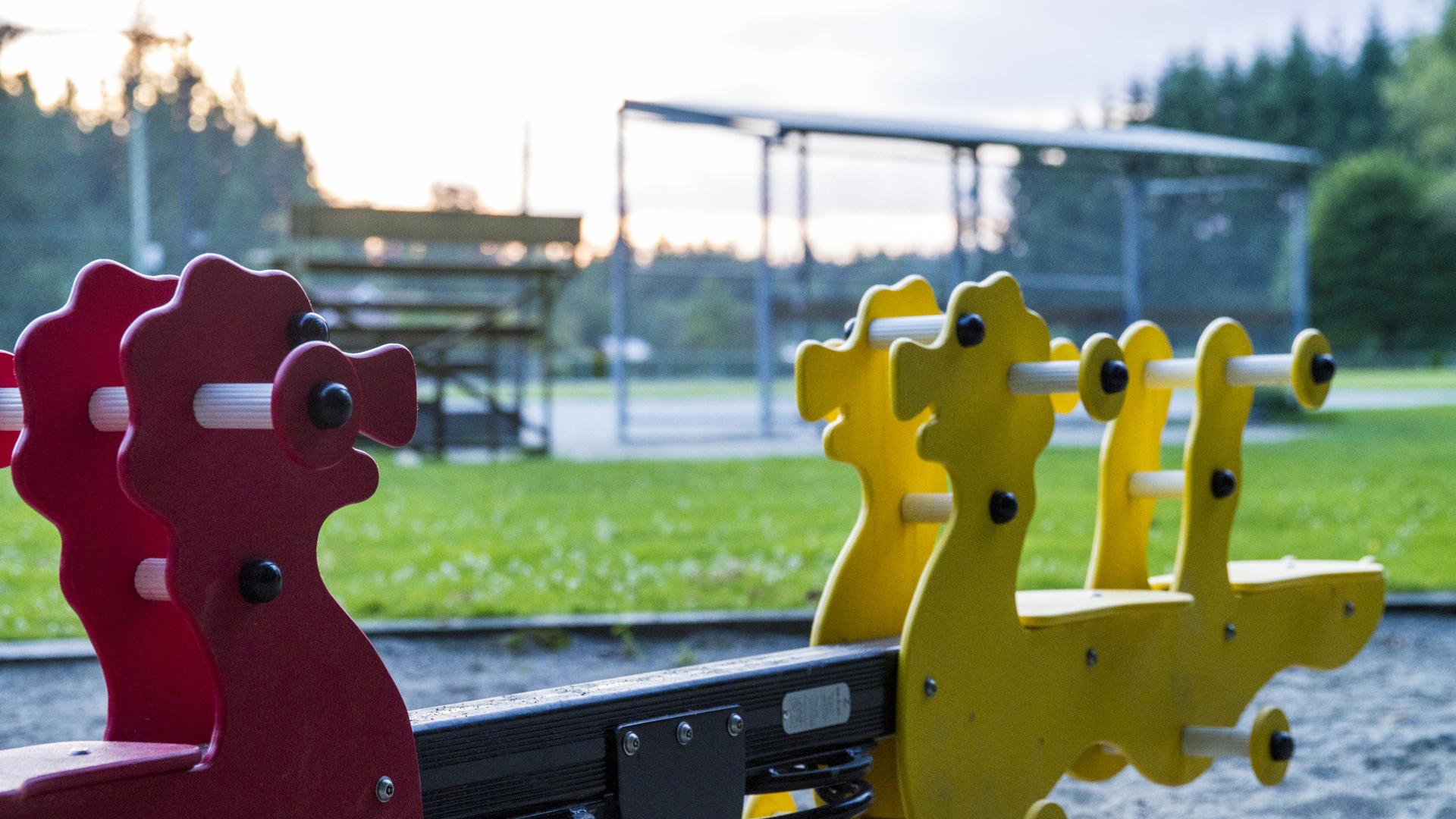 A red and yellow spring seesaw. In the background, a baseball diamond.