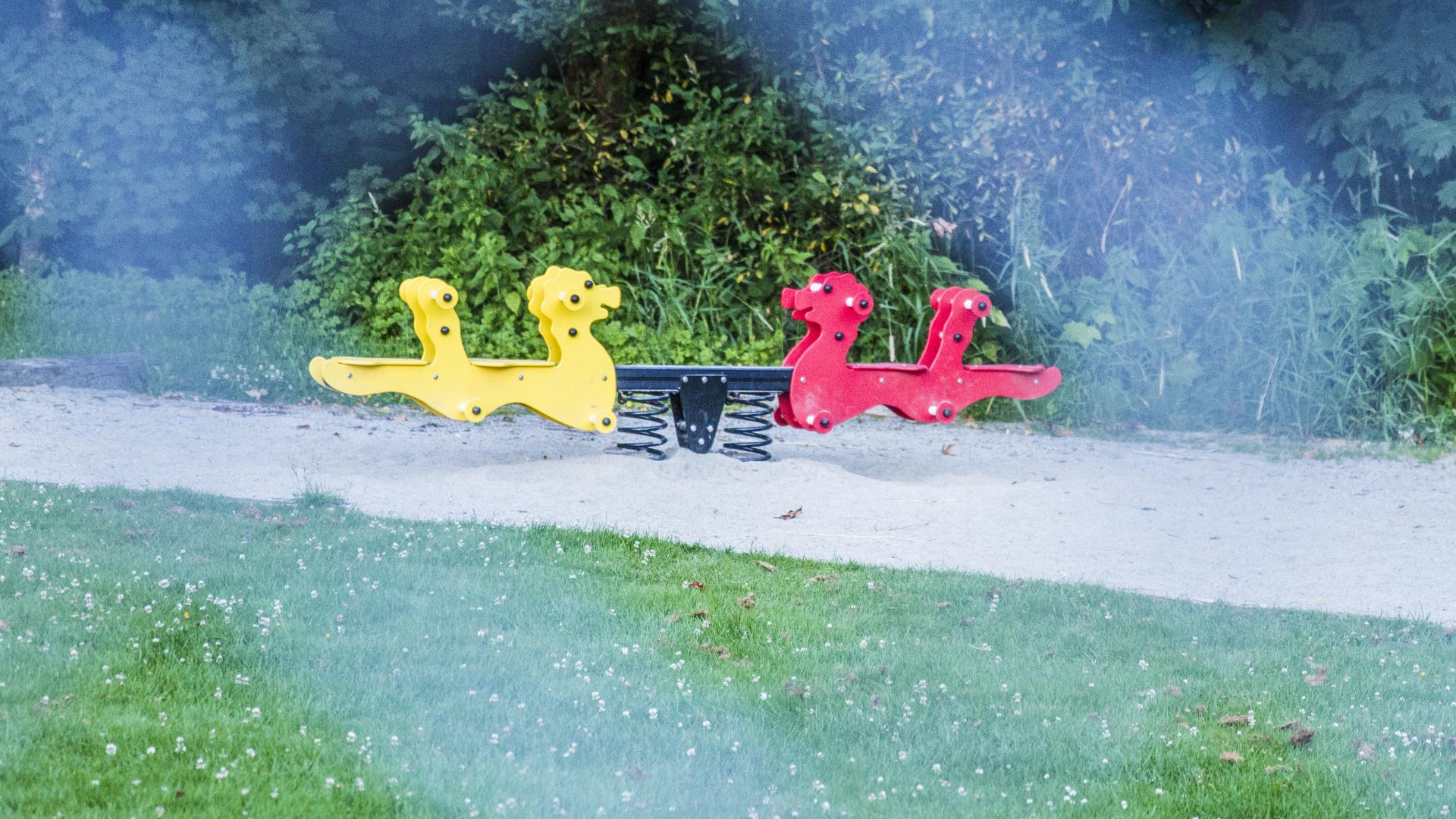 Through a chain link fence, a red and yellow spring seesaw. 