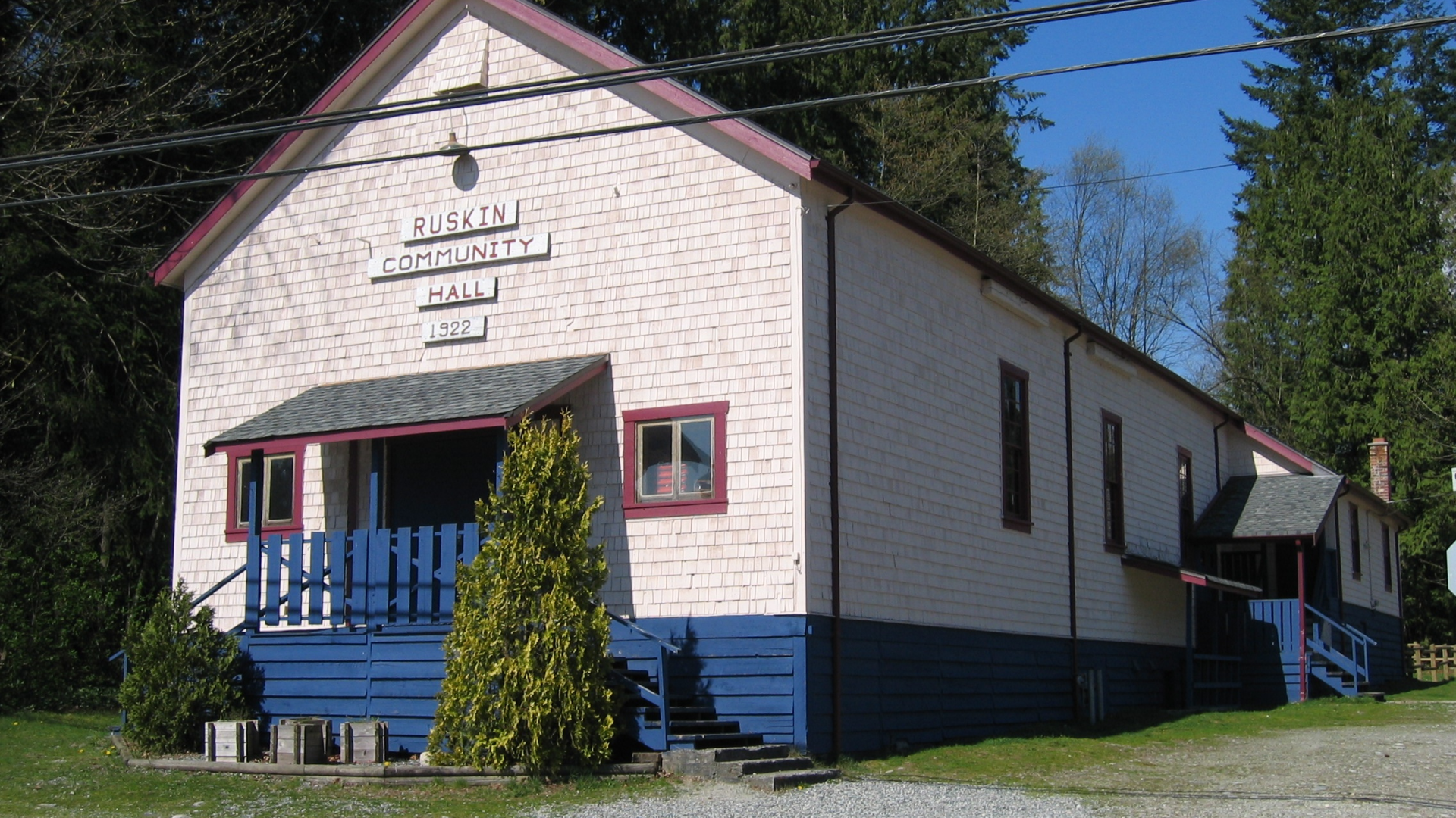 A blue and white painted building with a grey awning over the front door. The far side and rear of the building are surrounded by trees.