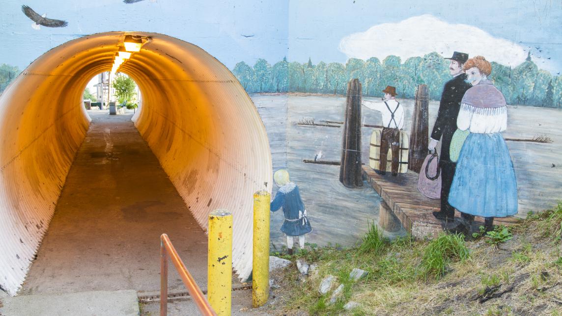 A mural of three people looking over the Fraser from a dock is painted on the outside of a round, covered walkway.