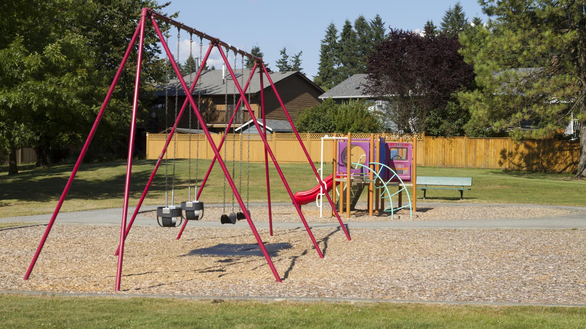 A swingset that is perpendicular to a paved path. On the other side of the path, there is a small playground.