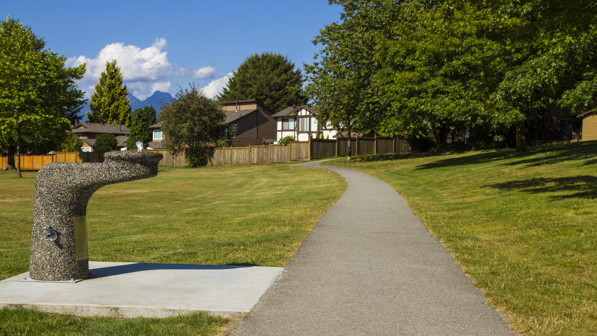 A stone water fountain on a foundation off a paved trail through an open field.