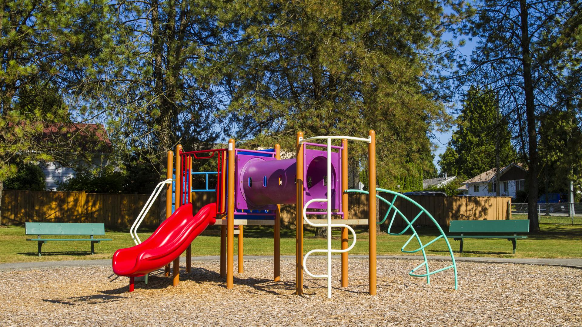 A small playground with some climbing equipment and a slide. Outside of the mulch bed, a bench is visible on either side of the playground.