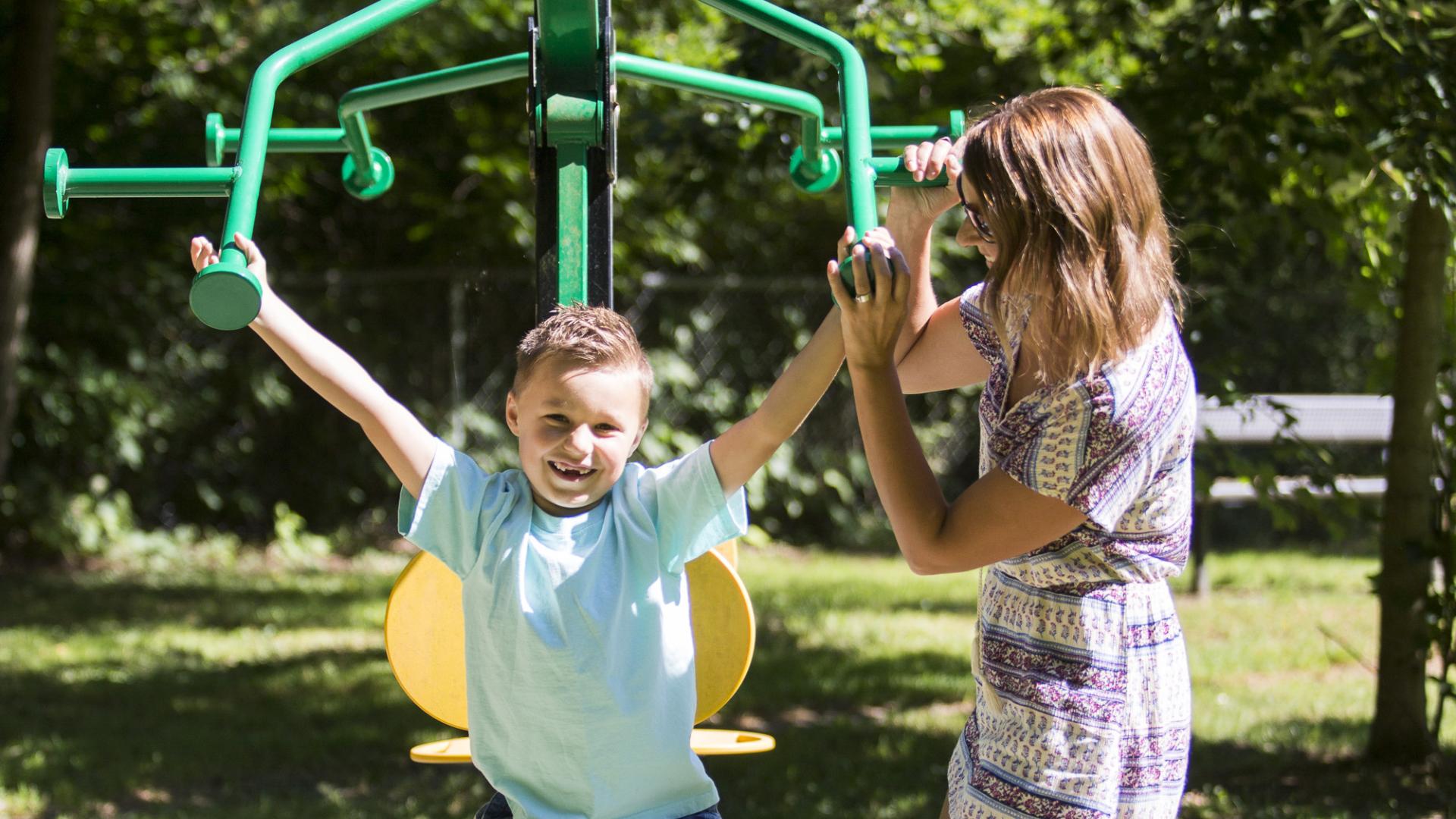 A pair of children use a piece of double sided manual park workout equipment. There is a seat on either side, and a cross beam that could be pulled down above the seats.