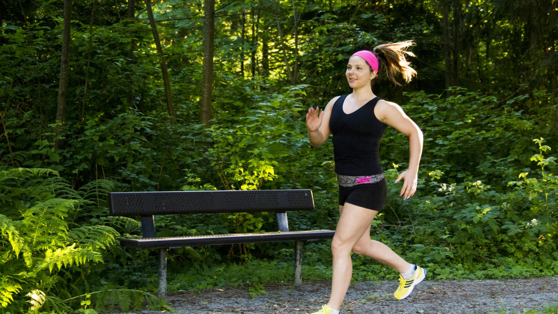A woman in workout clothes runs past a bench on a gravel trail, next to a forested area.