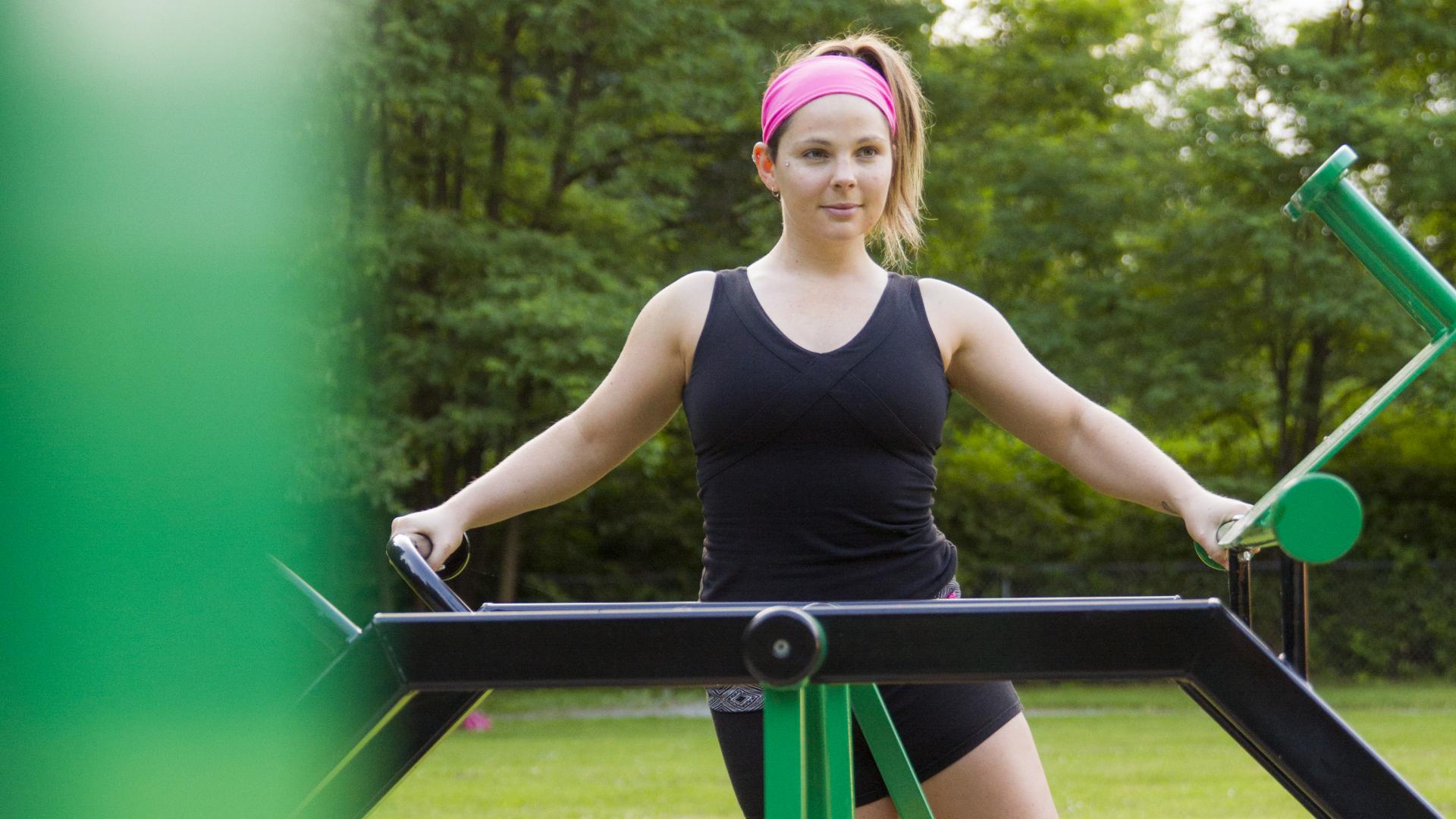 A women in workout gear uses a manual piece of park workout equipment. This one appears to swing back and forth as you stand on it.