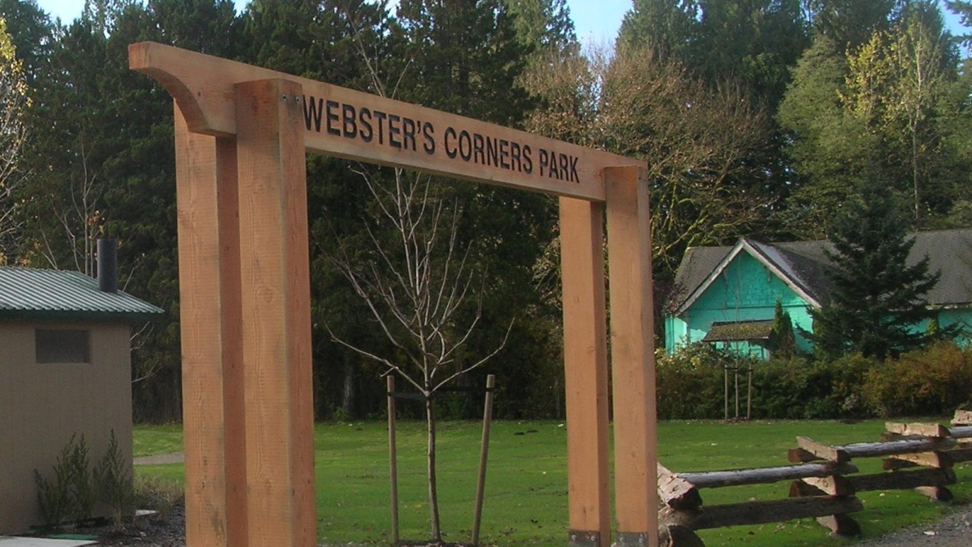 The Webster's Corner Park wooden archway sign over a wide sidewalk.
