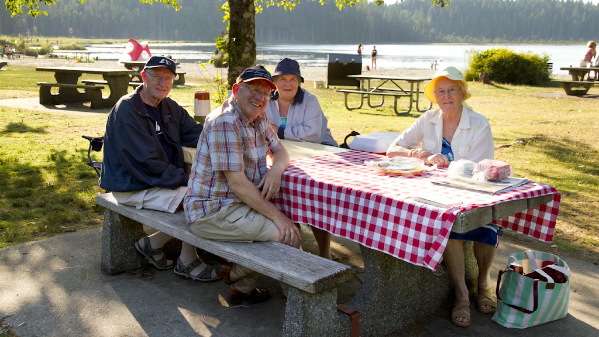Four adults sit and a picnic table near a large lake.