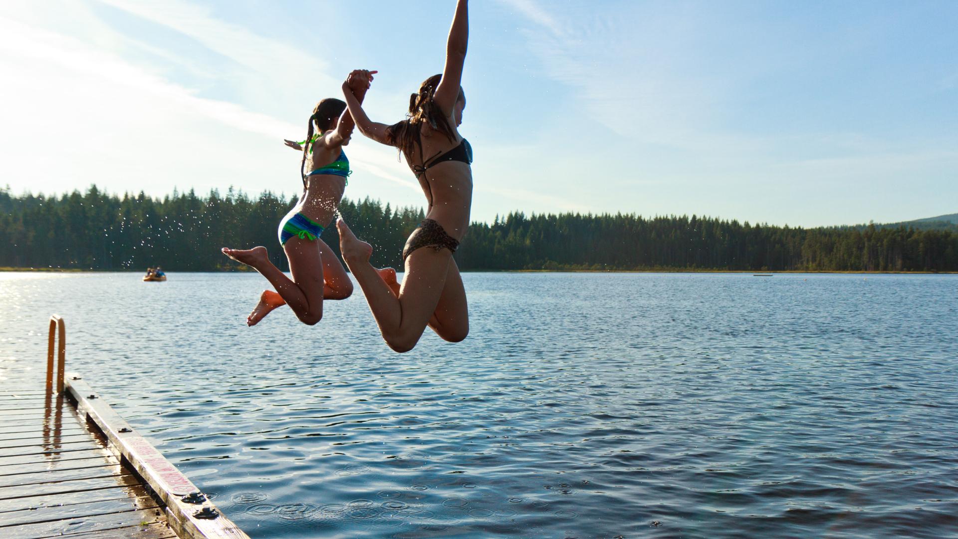 Two women jump off a dock with their hands in the air.