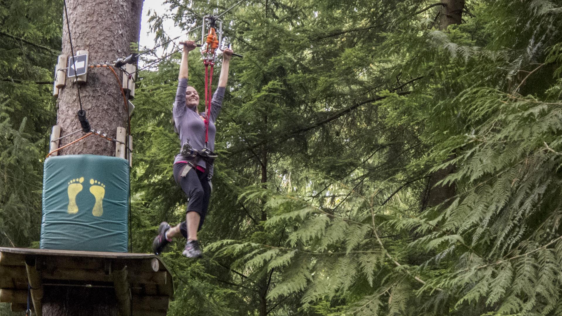 A woman leaps off a platform on a tree trunk. Her arms reach above her, appear to be grabbing a zip line. She is wearing a safety harness. 
