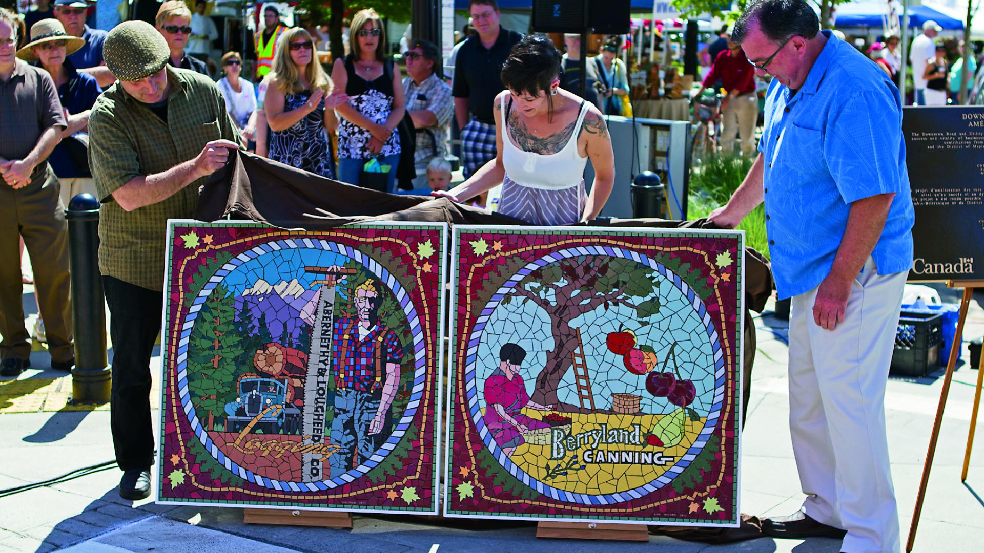 A crowd watches as three people display two tile mosaics. One depicts a man with logging equipment, while the other shows an androgynous worker sitting under a tree selecting and next to various fruit. White lettering proclaims "Berryland Canning."