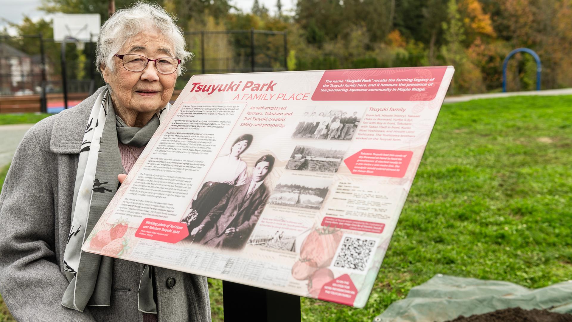 Tori Tsuyuki, an elderly woman and a member of the Tsuyuki family stands next to a plaque detailing their family and local heritage, which the park honors, with her wedding photo as the centrepiece.