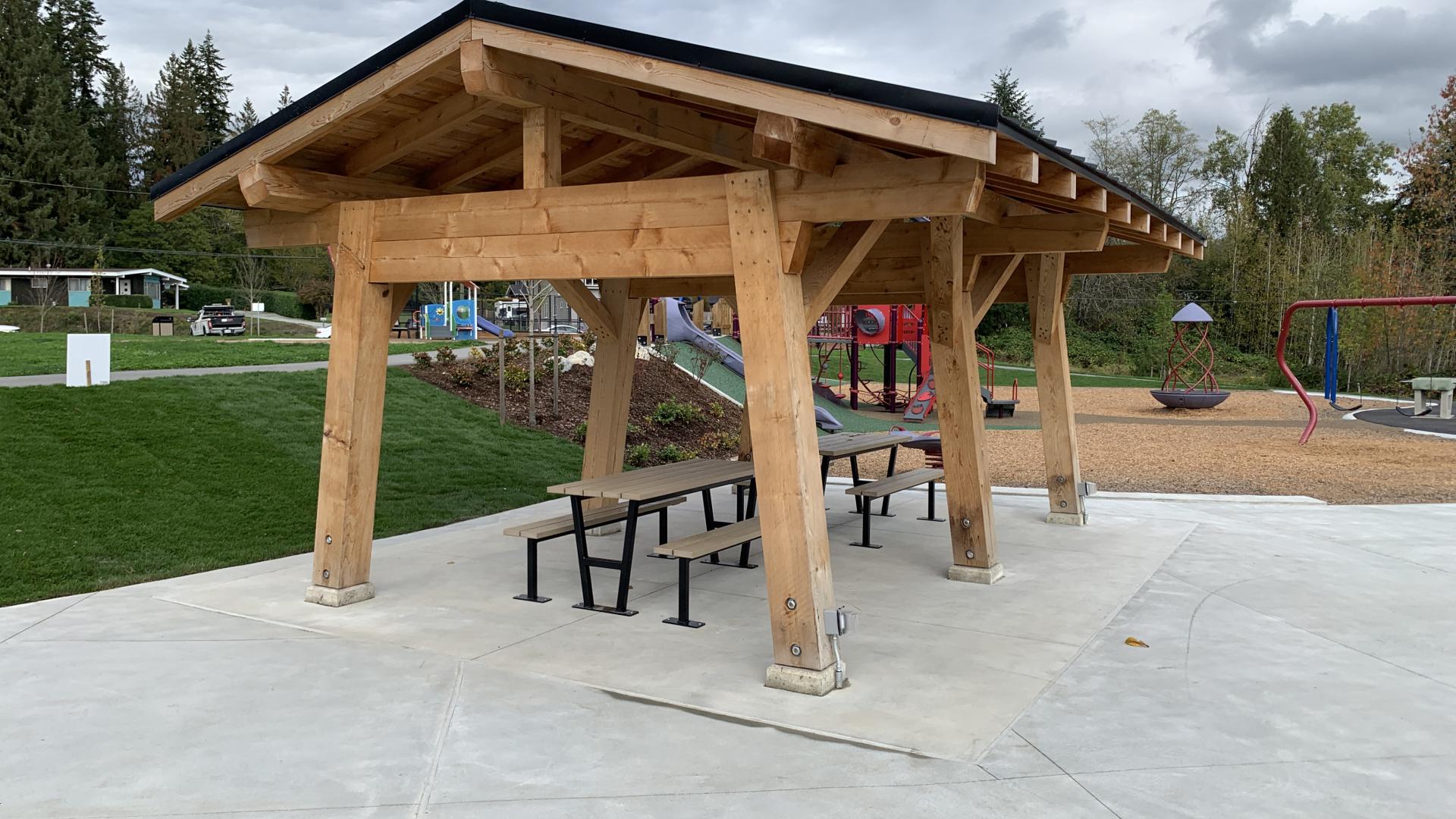 A wooden shelter covers two picnic tables overlooking the grass and playground of Tusyuki Park.