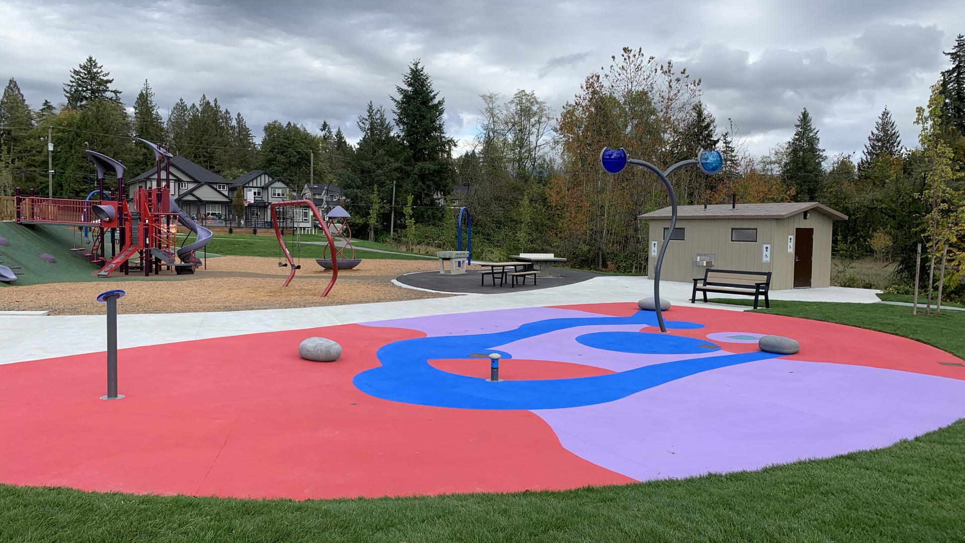 A purple, red and blue splash pad sits dormant between fresh grass and a paved pathway. In the background is a washroom building and play equipment.