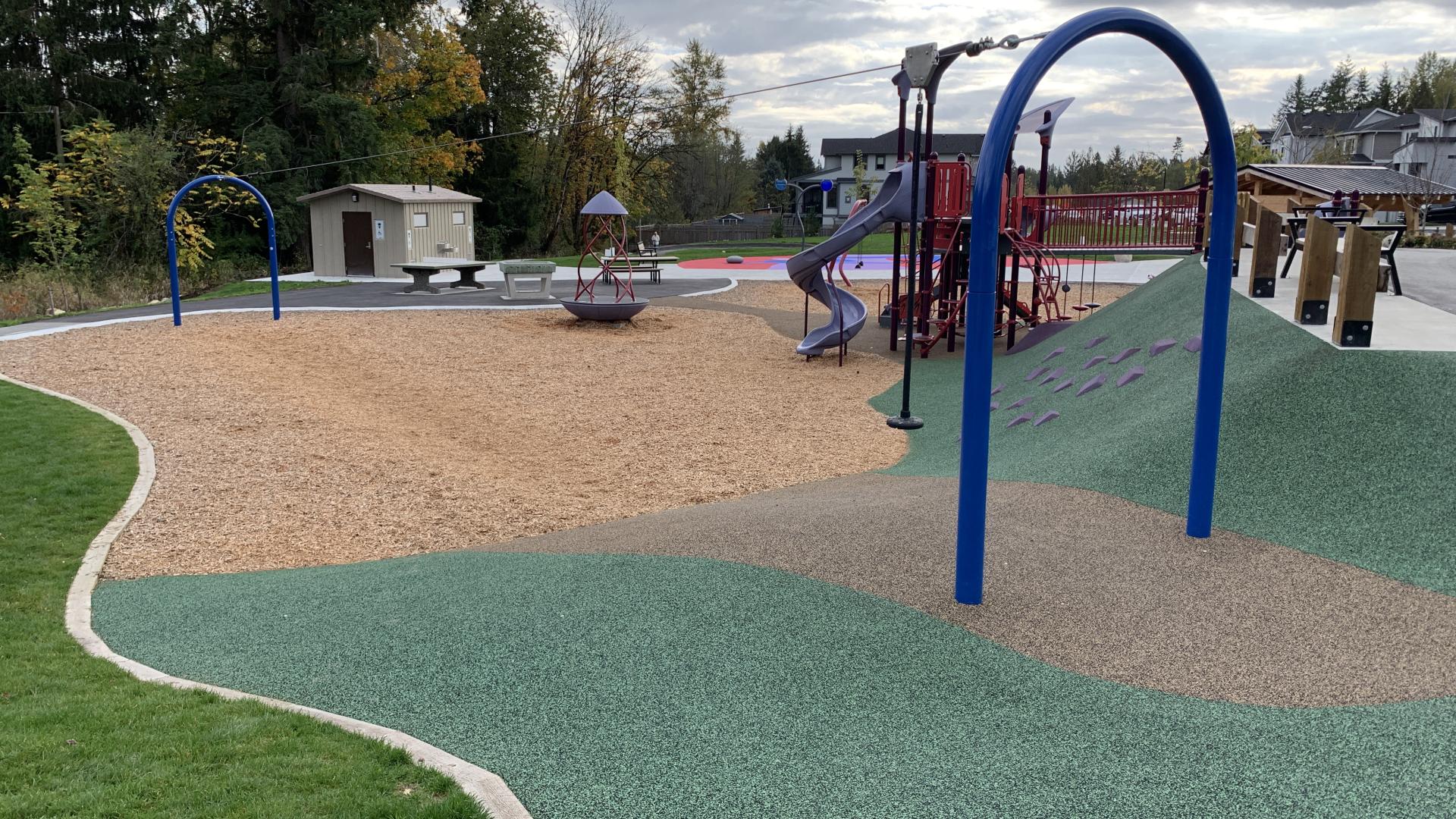 The play area is separated from the grass by a ridge of planks, and consists of a mix of bark mulch, rubber track/playground surfacing material, and paved areas. There is a zip line between two blue archways, and a red and blue playground in the background.