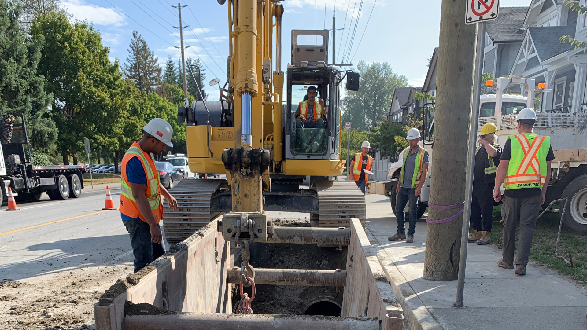 A crew of workers wearing high-visibility vests set a section of pipe into a construction canal next to the sidewalk.