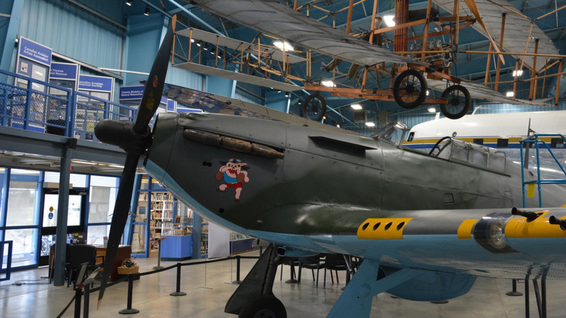 A plane with a nose propeller sits under a ceiling-mounted biplane in a museum exhibit.
