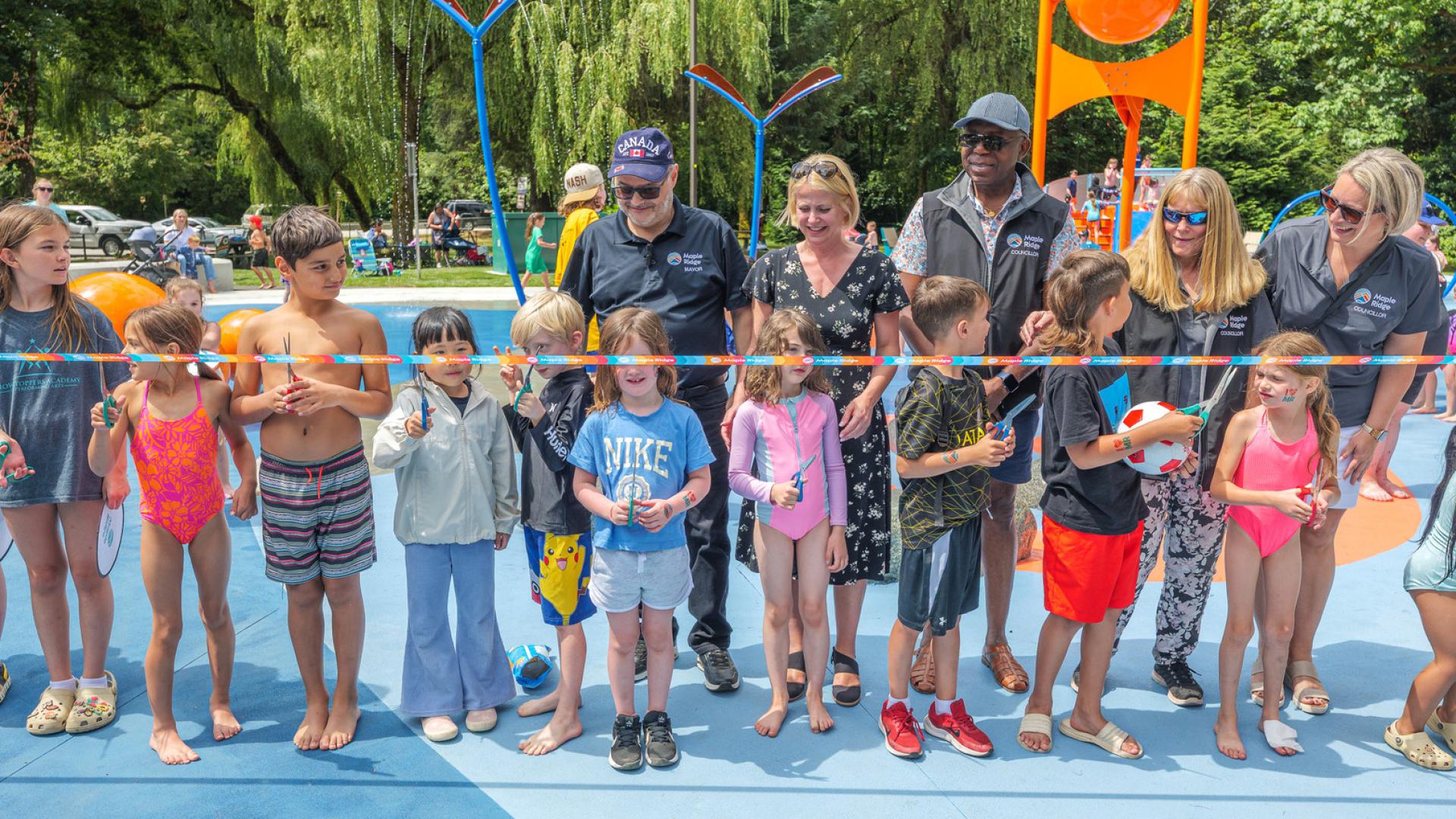 Mayor Dan Ruimy, MLA Lisa Beare, and Maple Ridge Councillors Judy Dueck, Korleen Carreras and Onyeka Dozie stand behind a line of kids, ready to cut the ribbon and officially open the Maple Ridge spray park.