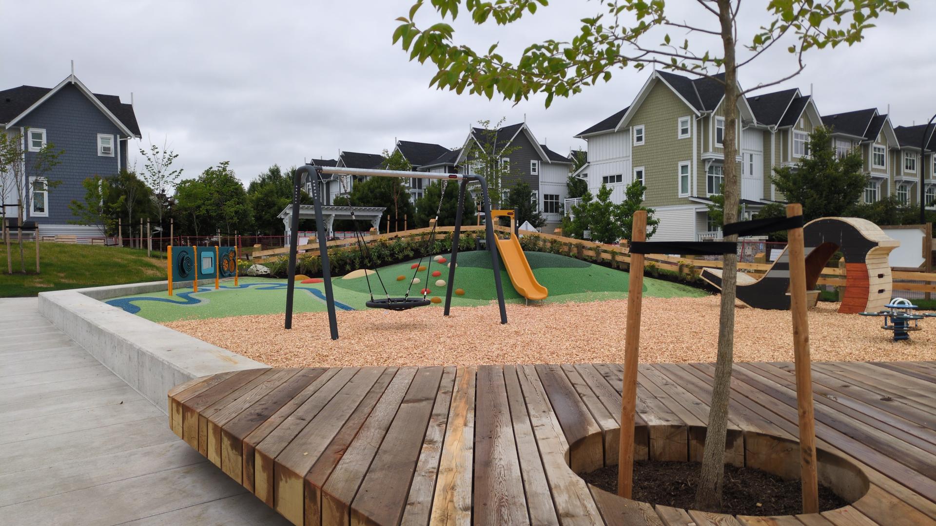 A heavy timber seating deck in a concrete plaza with playground behind with hill slide and disc swing