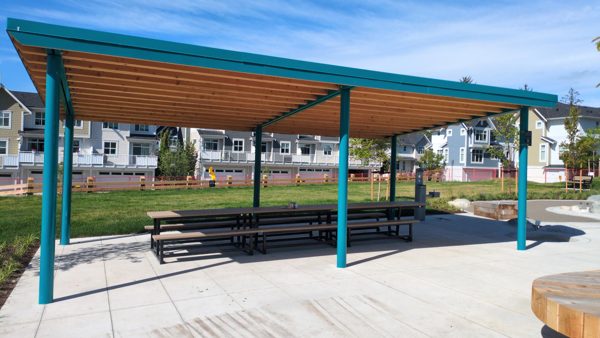 A teal metal picnic shelter with 3 picnic tables in front of open lawn space in a concrete plaza