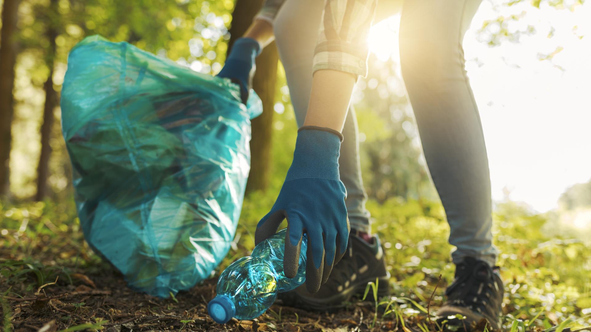 A woman bends down to pick up a water bottle off the forest floor to add to her blue bag of trash.
