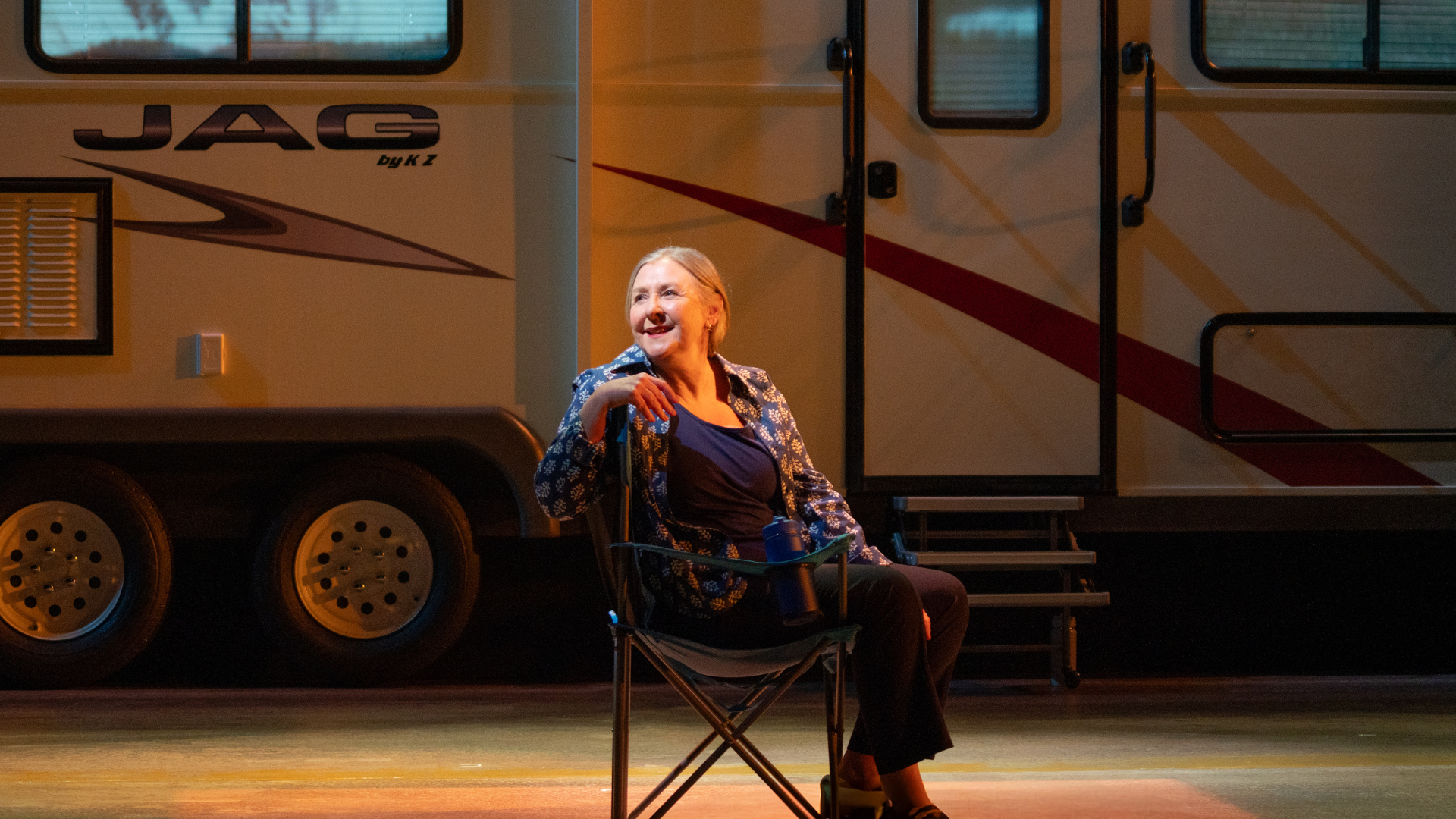A female performer sits onstage in a camping chair outside a recreational vehicle.