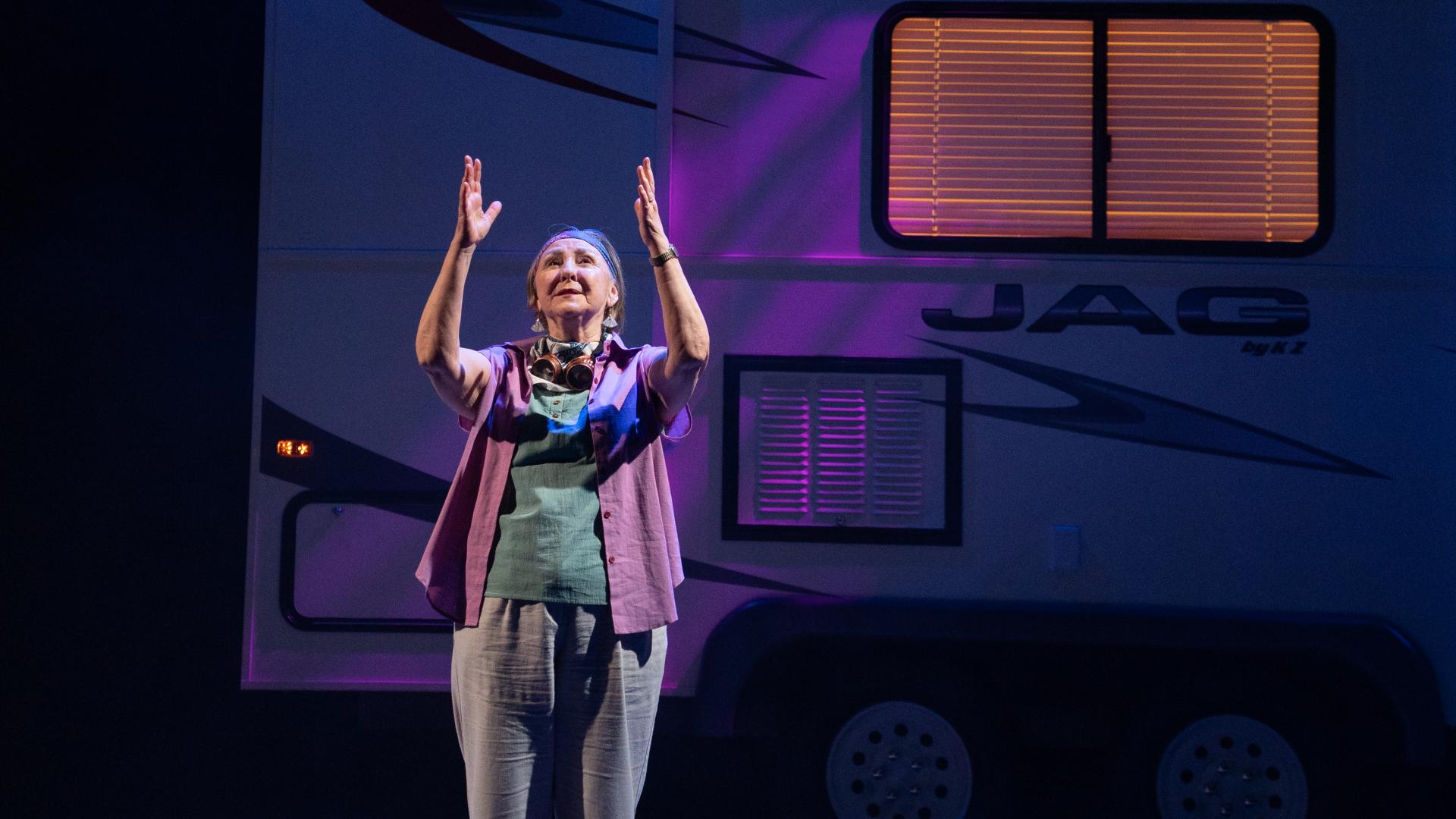 A female performer stands with her hands upwards onstage in a dimly lit scene in front of a recreational vehicle.